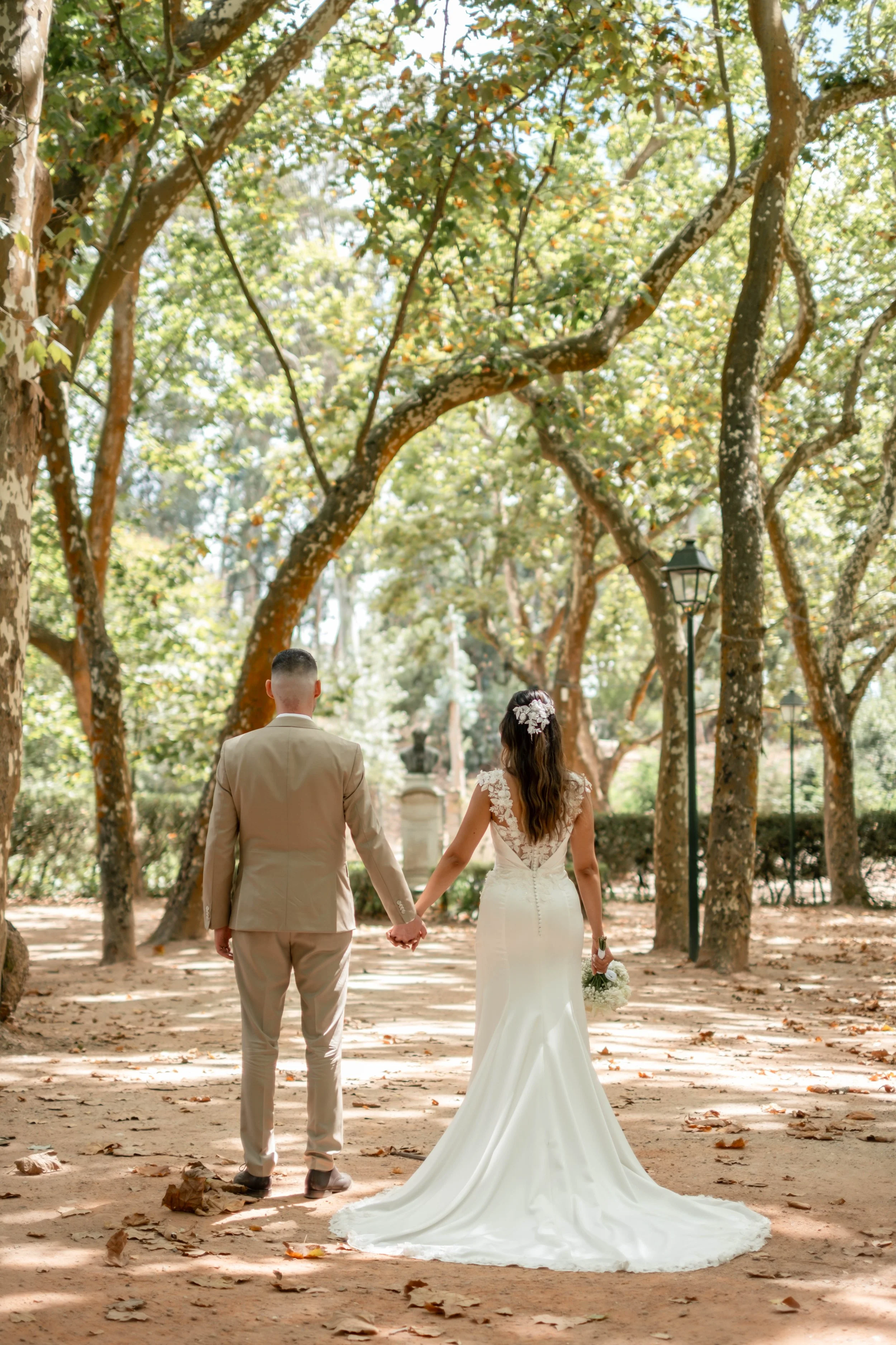 Noivos de mãos dadas vistos de costas num caminho arborizado