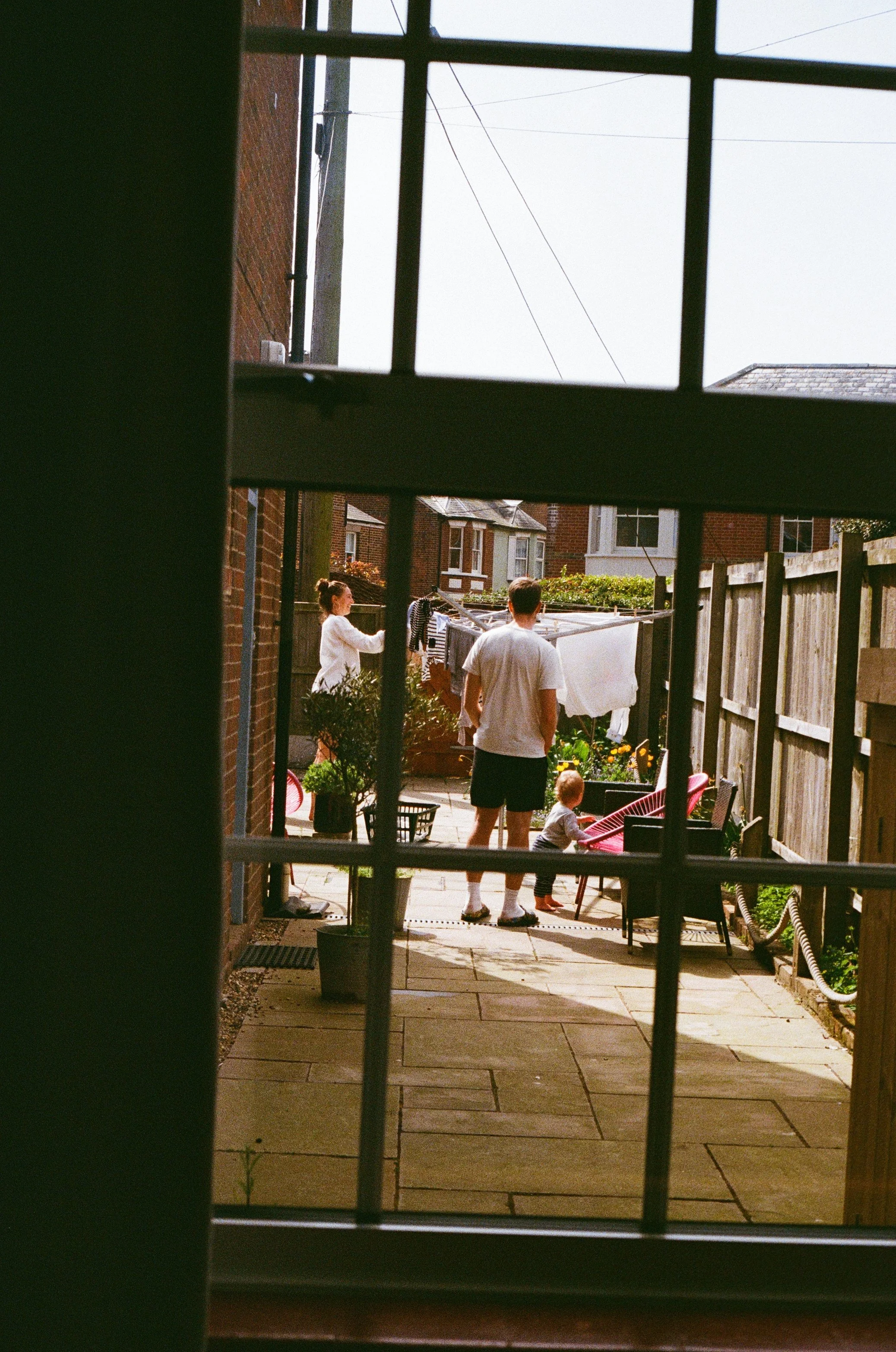 Through a windowpane you can see a man and woman hanging out washing whilst their toddler explores the garden.