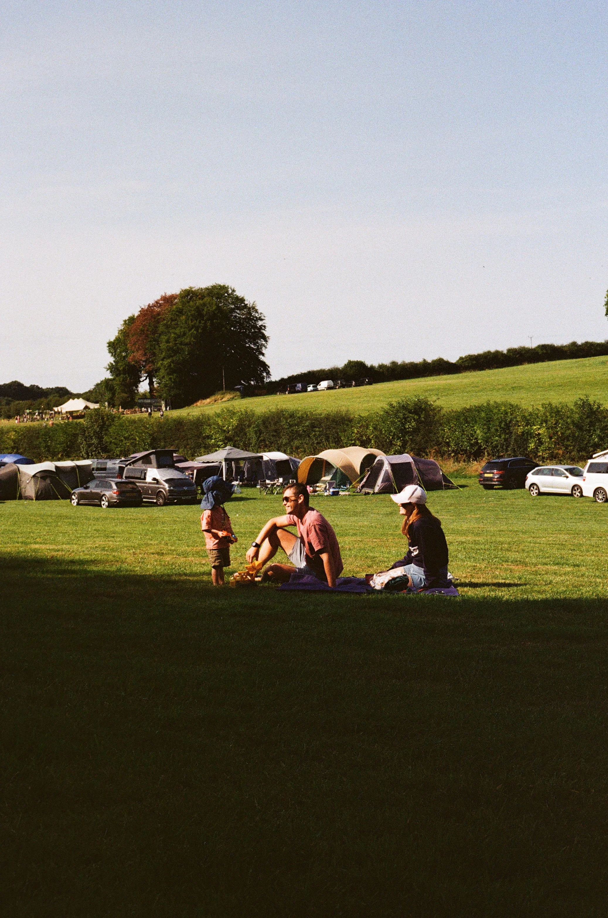 Family camping scene with tents, parked cars, and green fields under a clear sky.