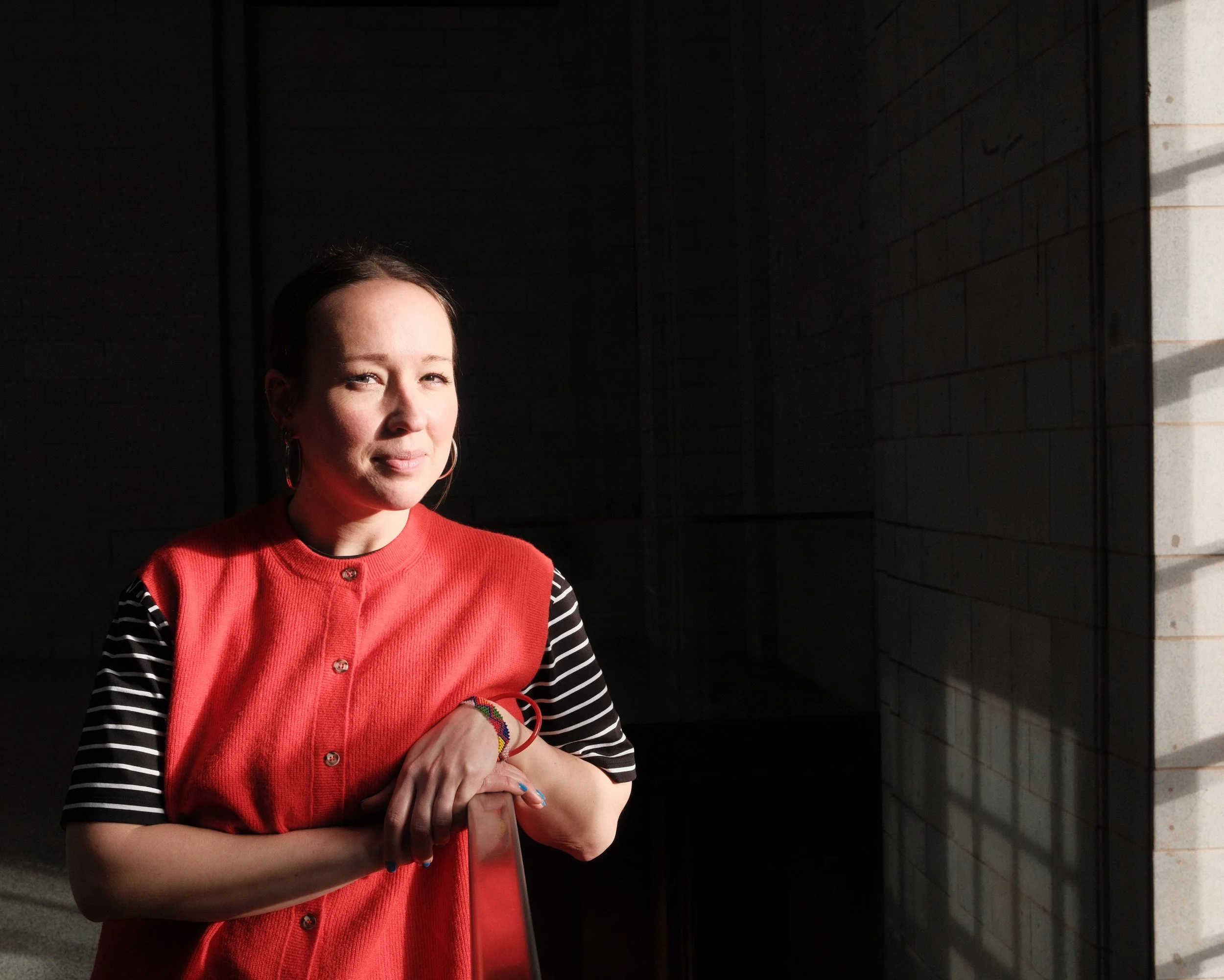 A woman with light skin, dark hair, and hoop earrings, dressed in a red vest over a black and white striped shirt, standing in front of a dark background with sunlight shining on her face and right arm.