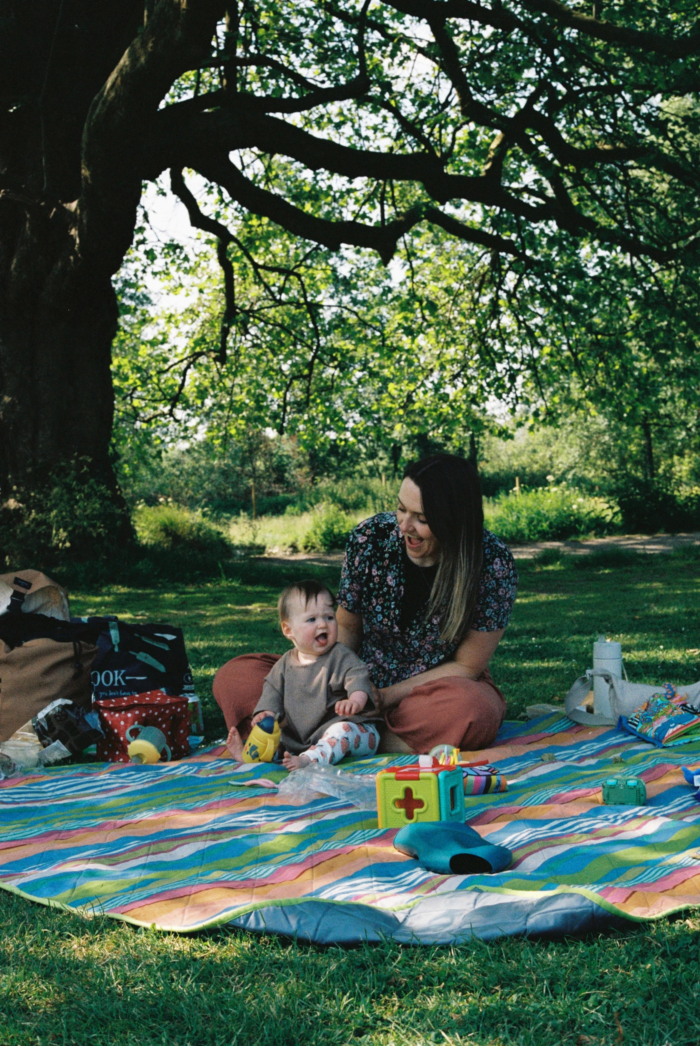 A woman and a baby sitting on a colorful blanket under a large tree, surrounded by toys and picnic supplies in a park.