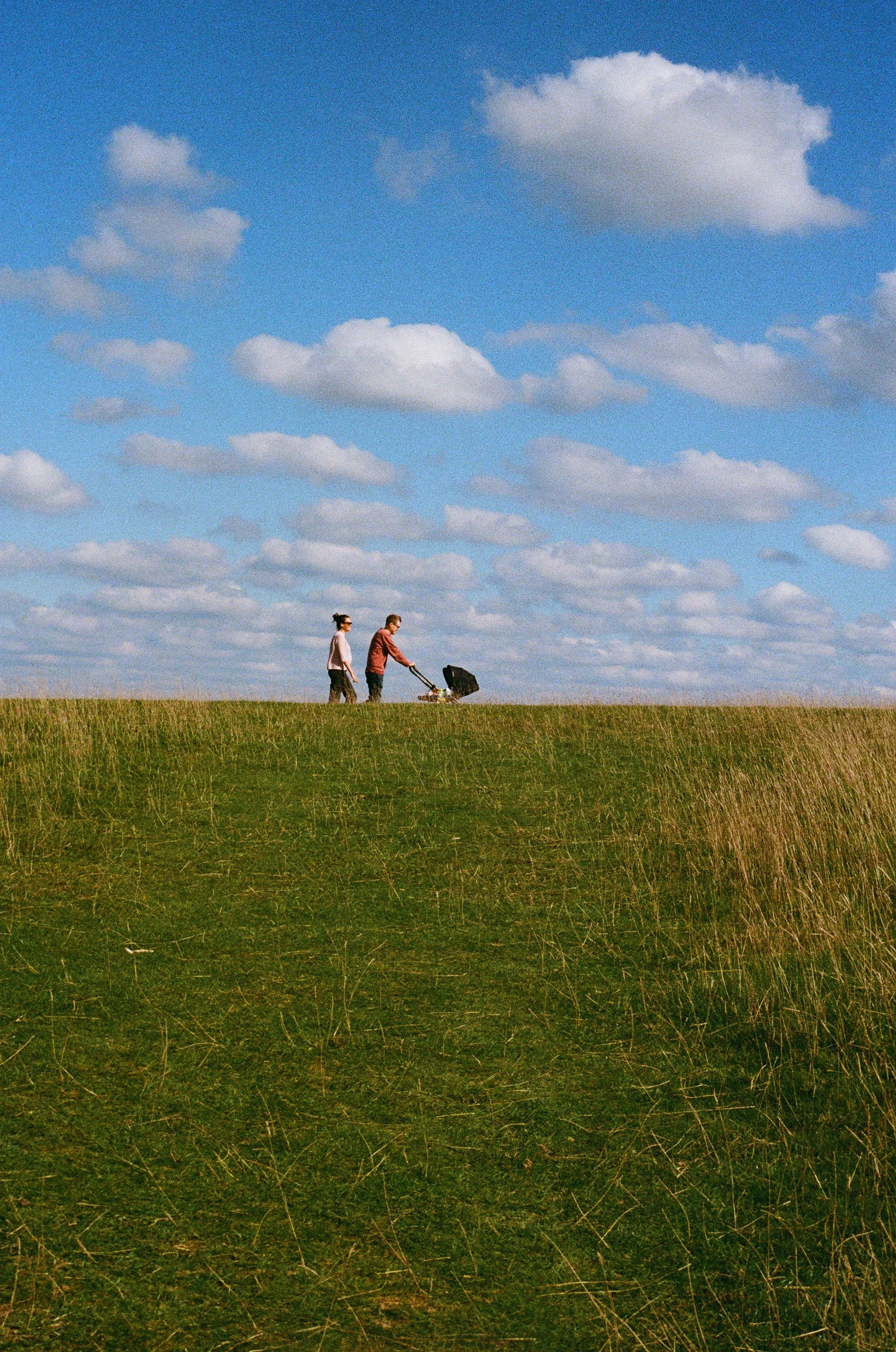 A man pushing a pram on the top of a grassy hill with a woman walking beside him under a blue sky with scattered clouds.