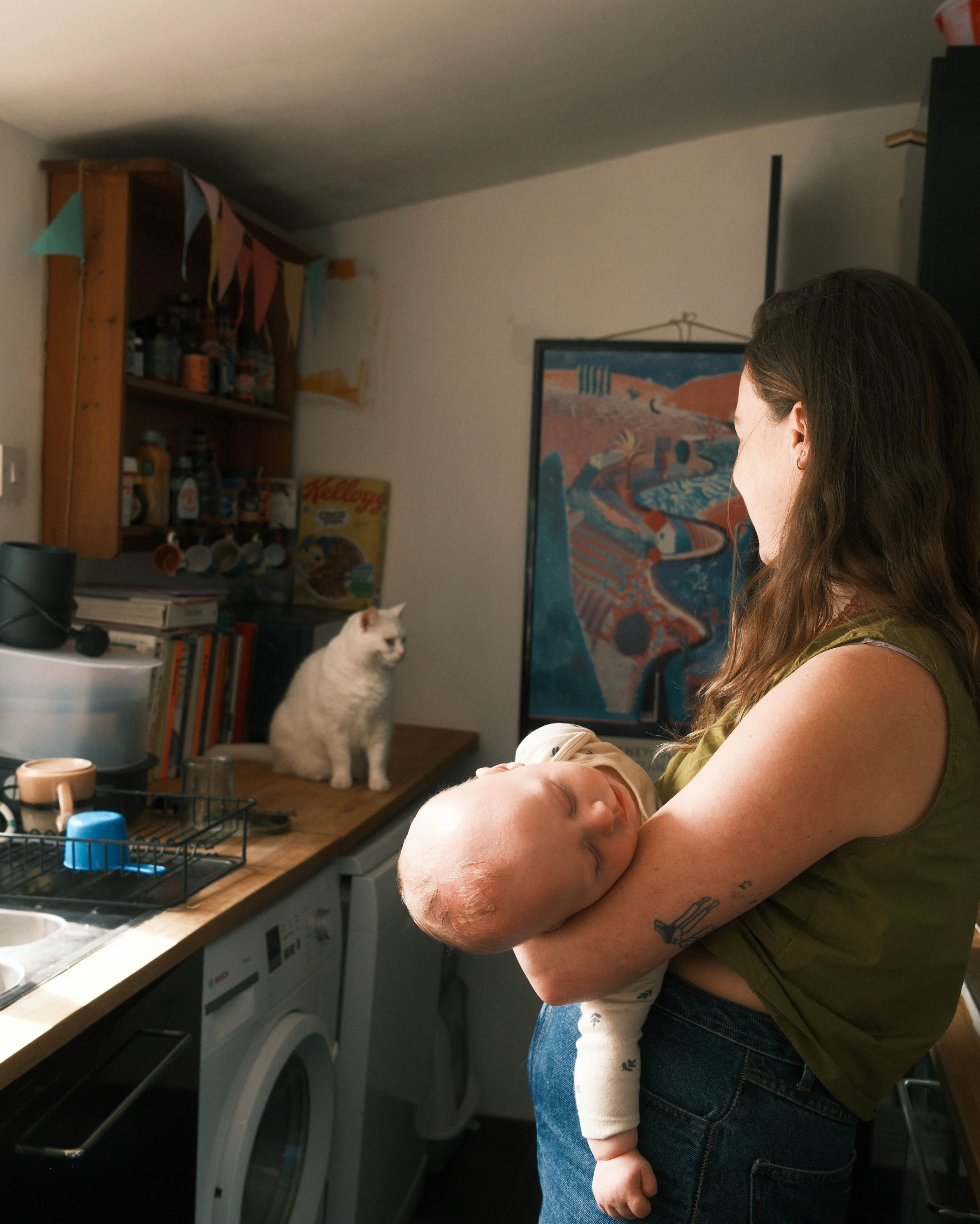 A mother holds her sleeping baby in her arms in her kitchen. She is standing side on and looking towards her white cat, who is sat on the kitchen counter.