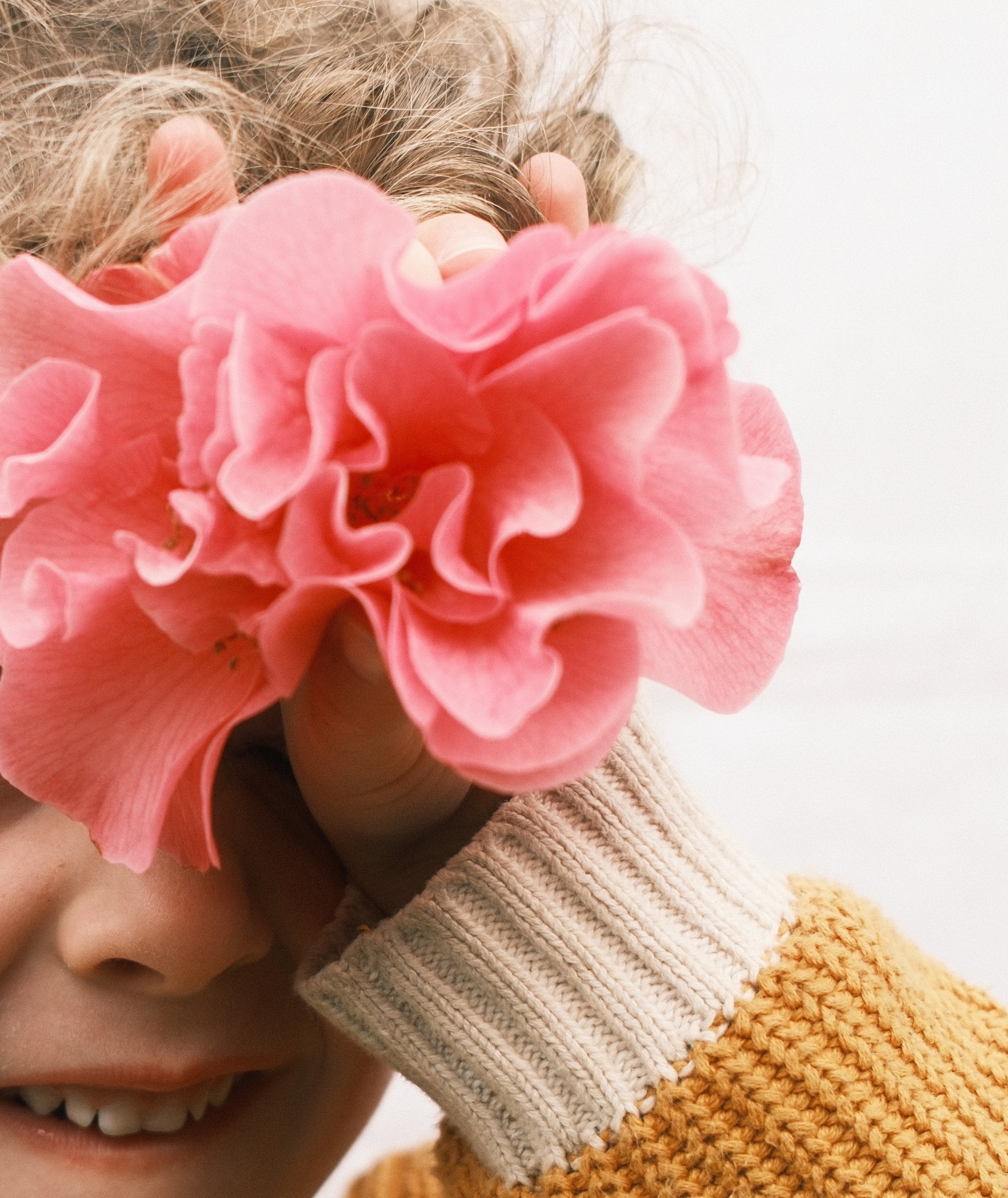 A person holding a pink flower up to their face, partially covering it. The person has light-colored hair, and is wearing a beige and yellow knit sweater.