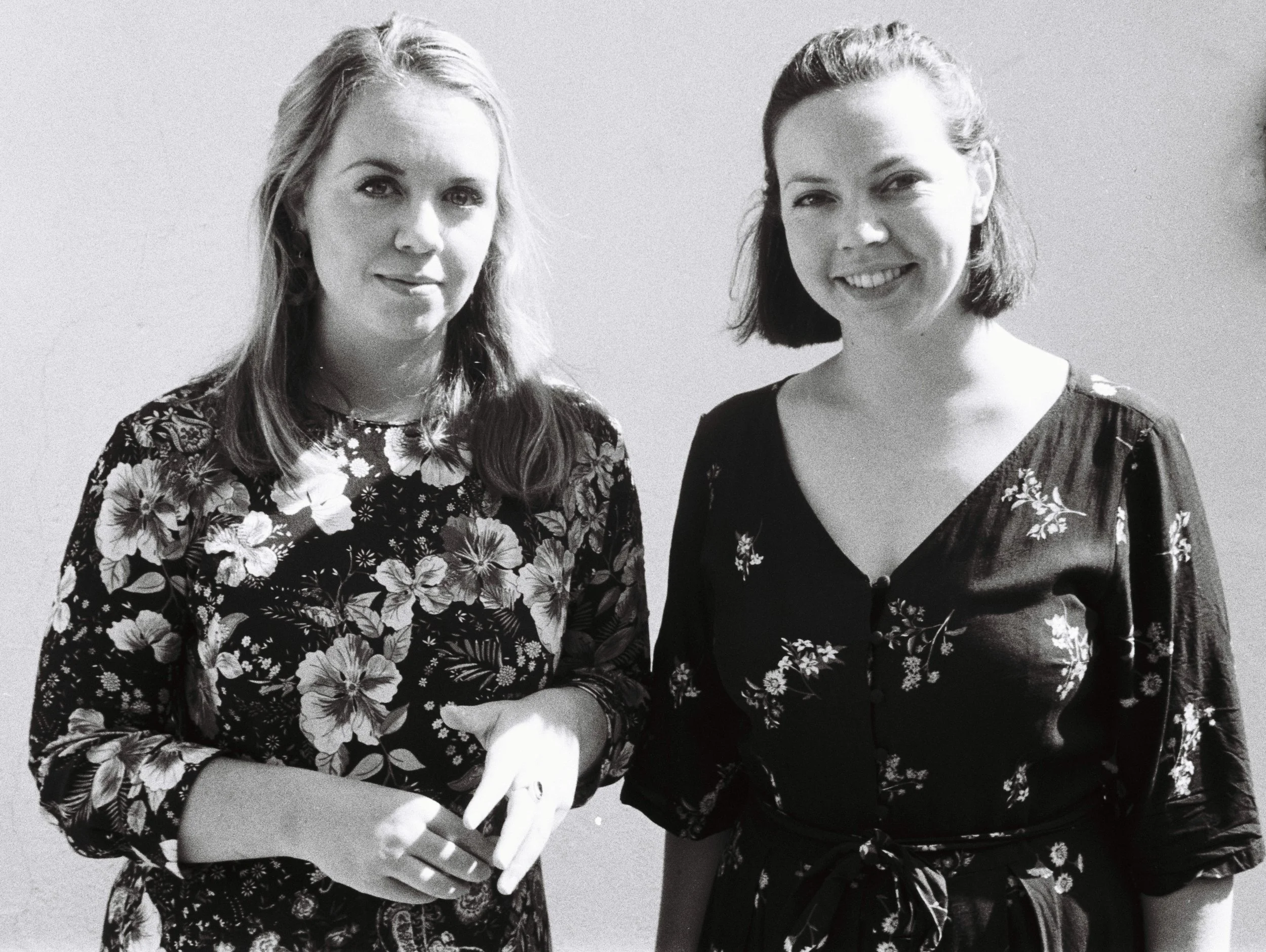 Two women standing side by side, both wearing floral patterned tops, smiling and looking at the camera, against a plain background.