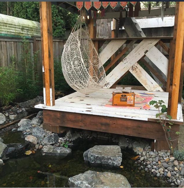 A small outdoor deck with a hanging white wicker egg chair, a vintage-style wooden radio, and a colorful rug. The deck is elevated over a small rocky stream with water and stones below.