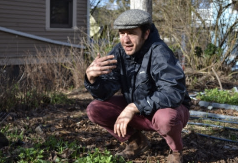 A man wearing a gray flat cap, black rain jacket, and maroon pants teaching in a garden during the day, gesturing and talking.