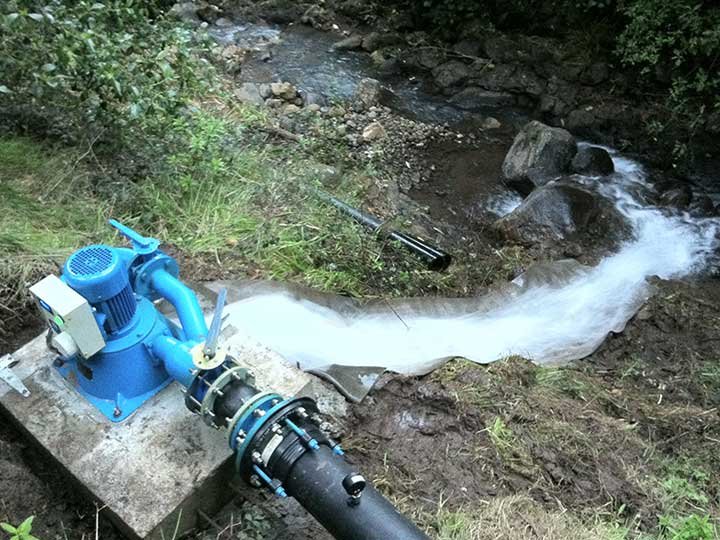 Water pump by a stream with flowing water and rocks in a natural outdoor setting.