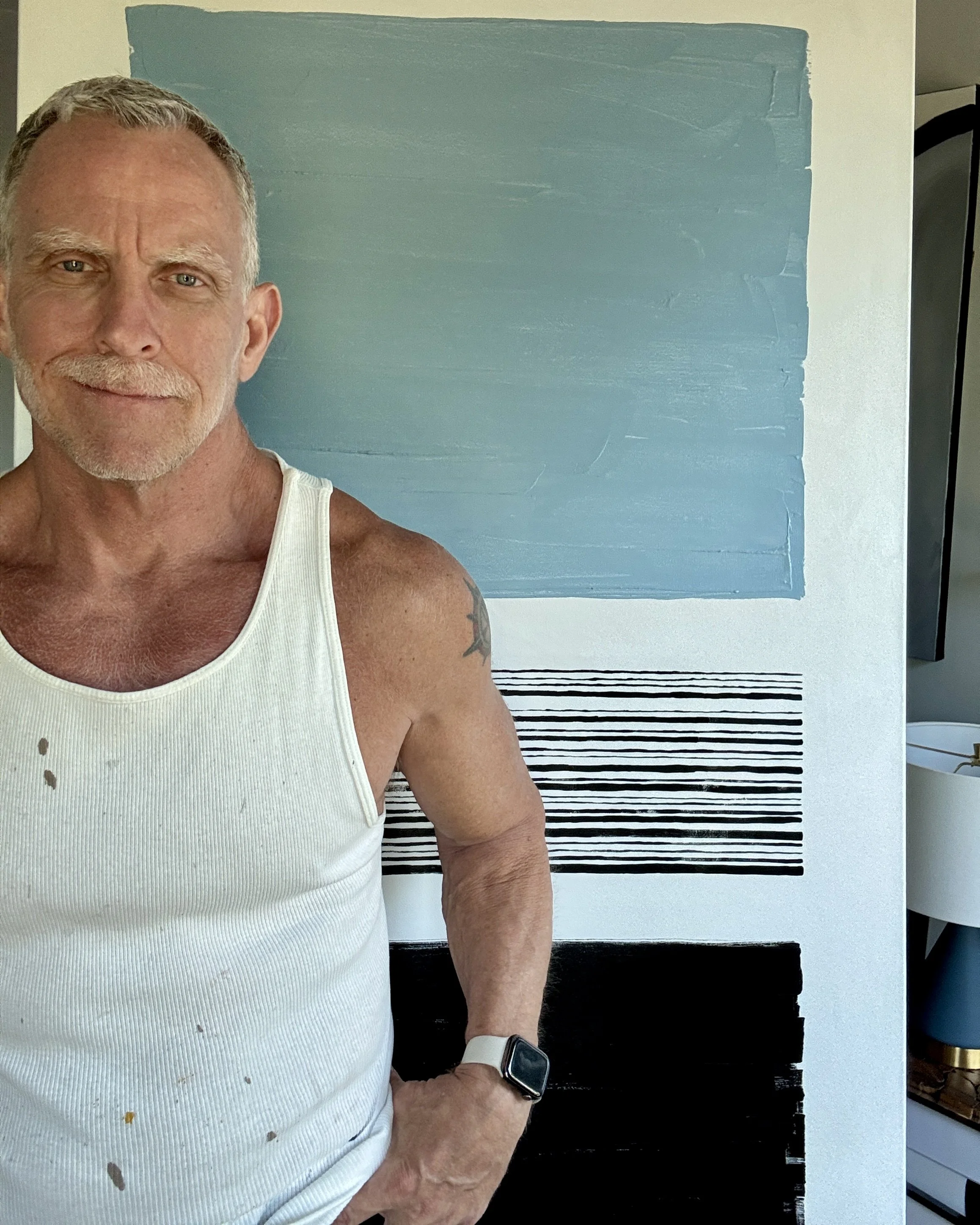 Close-up of an older man wearing a white tank top, a smartwatch, and a white wristband, standing in front of artwork with shades of blue and black stripes. His face and part of his arm are visible.