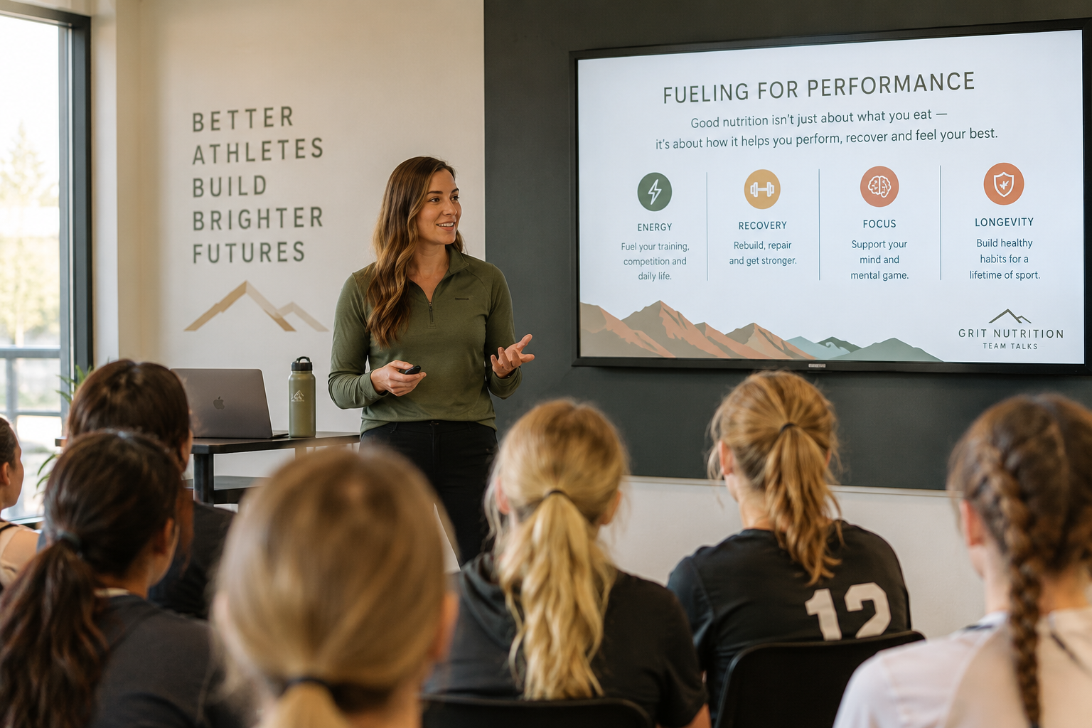 A woman giving a presentation to a group of young female athletes in a room with a mountain themed banner that says "Better Athletes Build Brighter Futures". The presentation slide discusses fueling for performance, including energy, recovery, focus, and longevity.