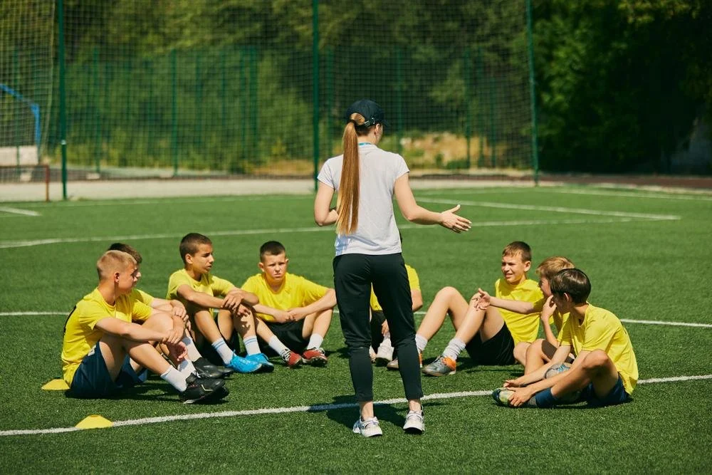 A coach or instructor giving instructions to a group of young boys in yellow sports uniforms sitting on a soccer field.