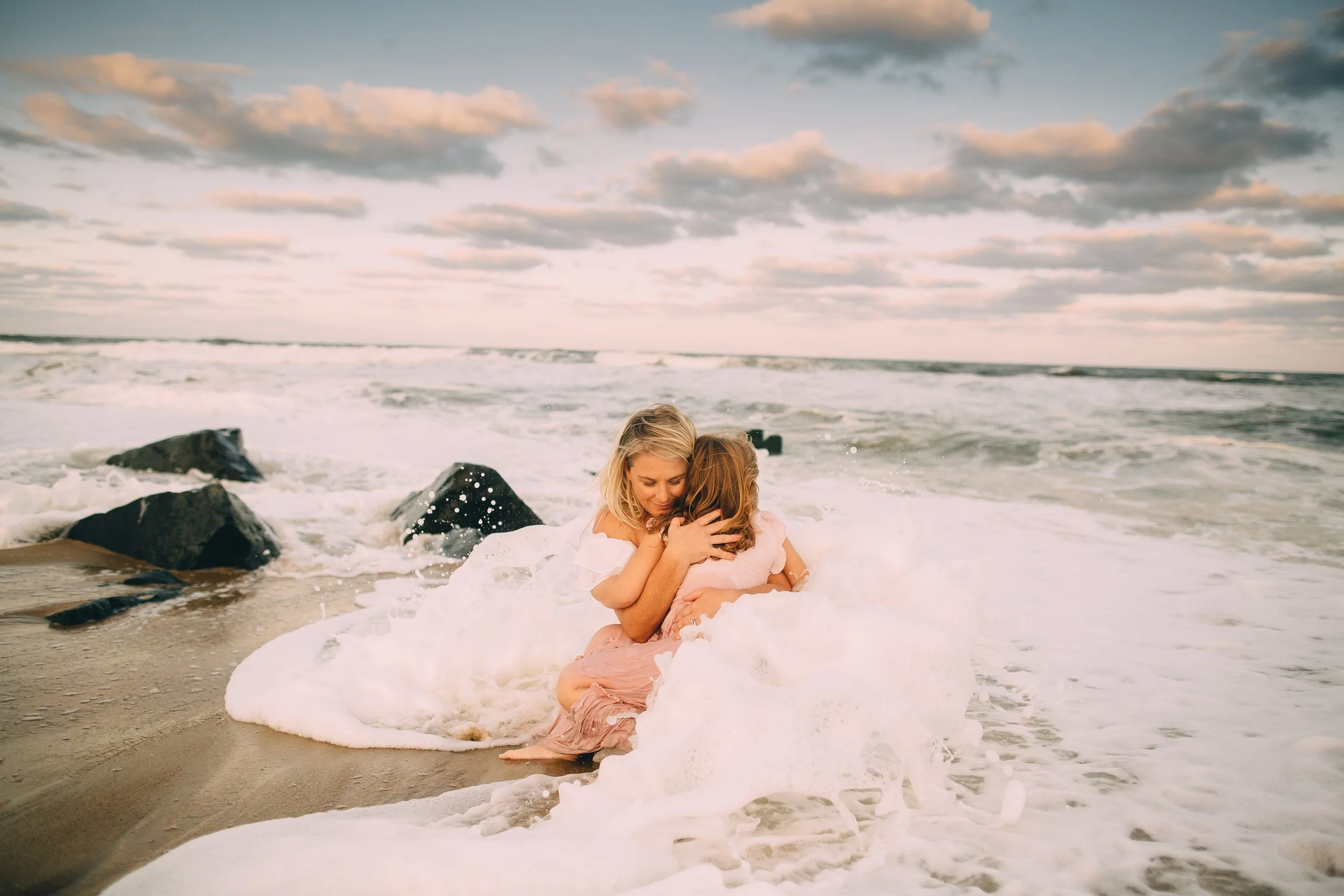 Mom and daughter  hugging by the ocean with waves and rocks in the background during sunset.