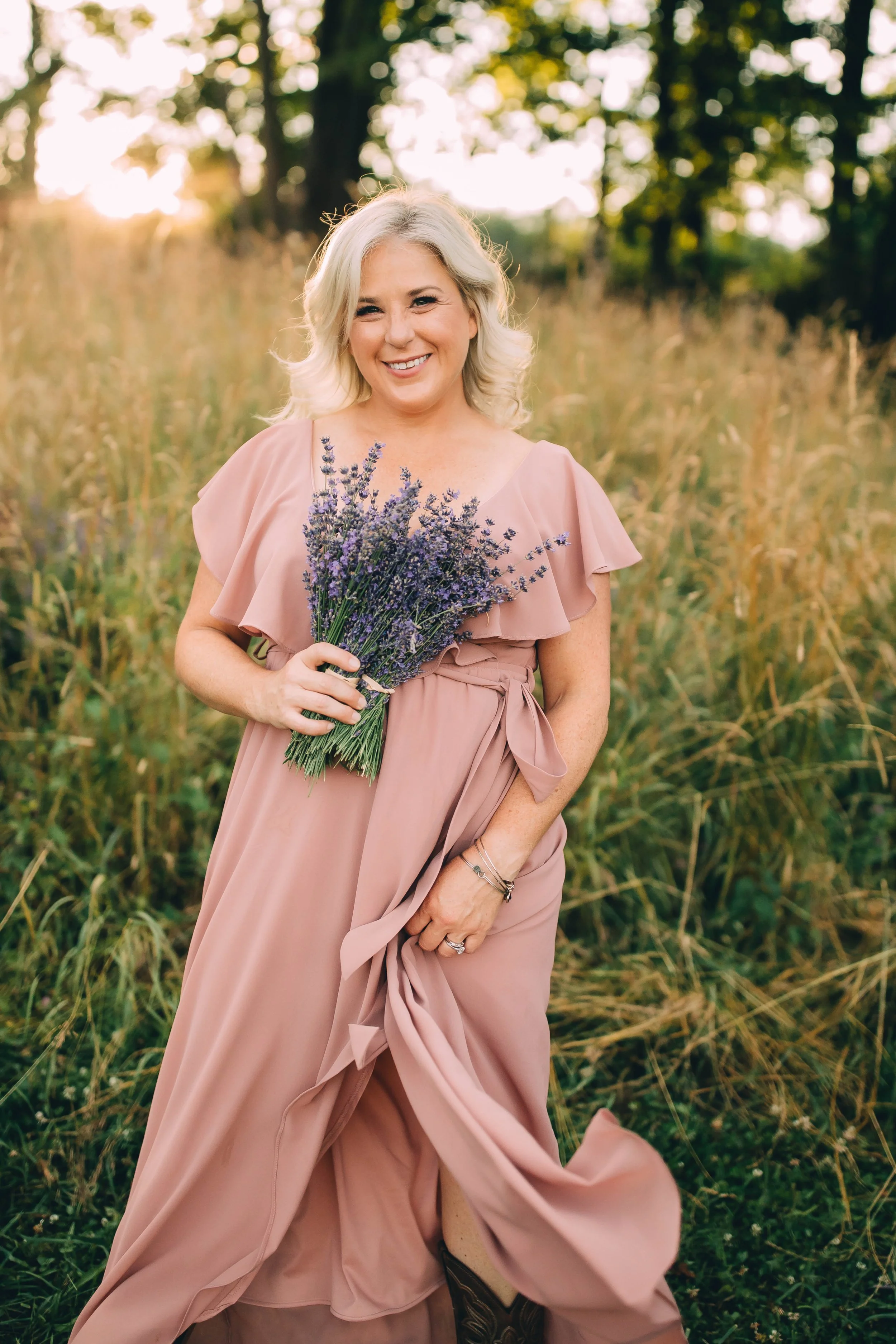 A smiling woman with blonde hair holding a bouquet of lavender flowers in a field of tall grass during sunset.