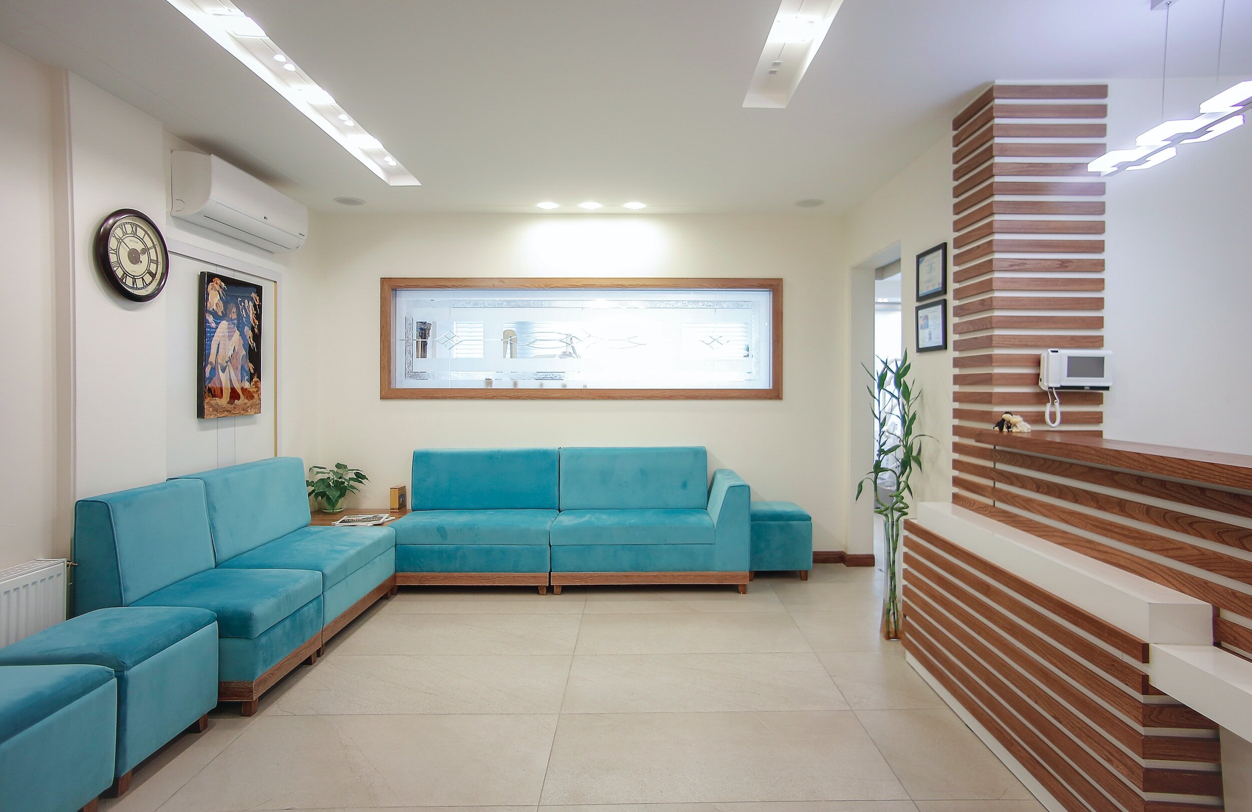 A modern waiting room with a teal sectional sofa, wall art, a wall clock, and a window with frosted glass, decorated with plants and framed pictures.
