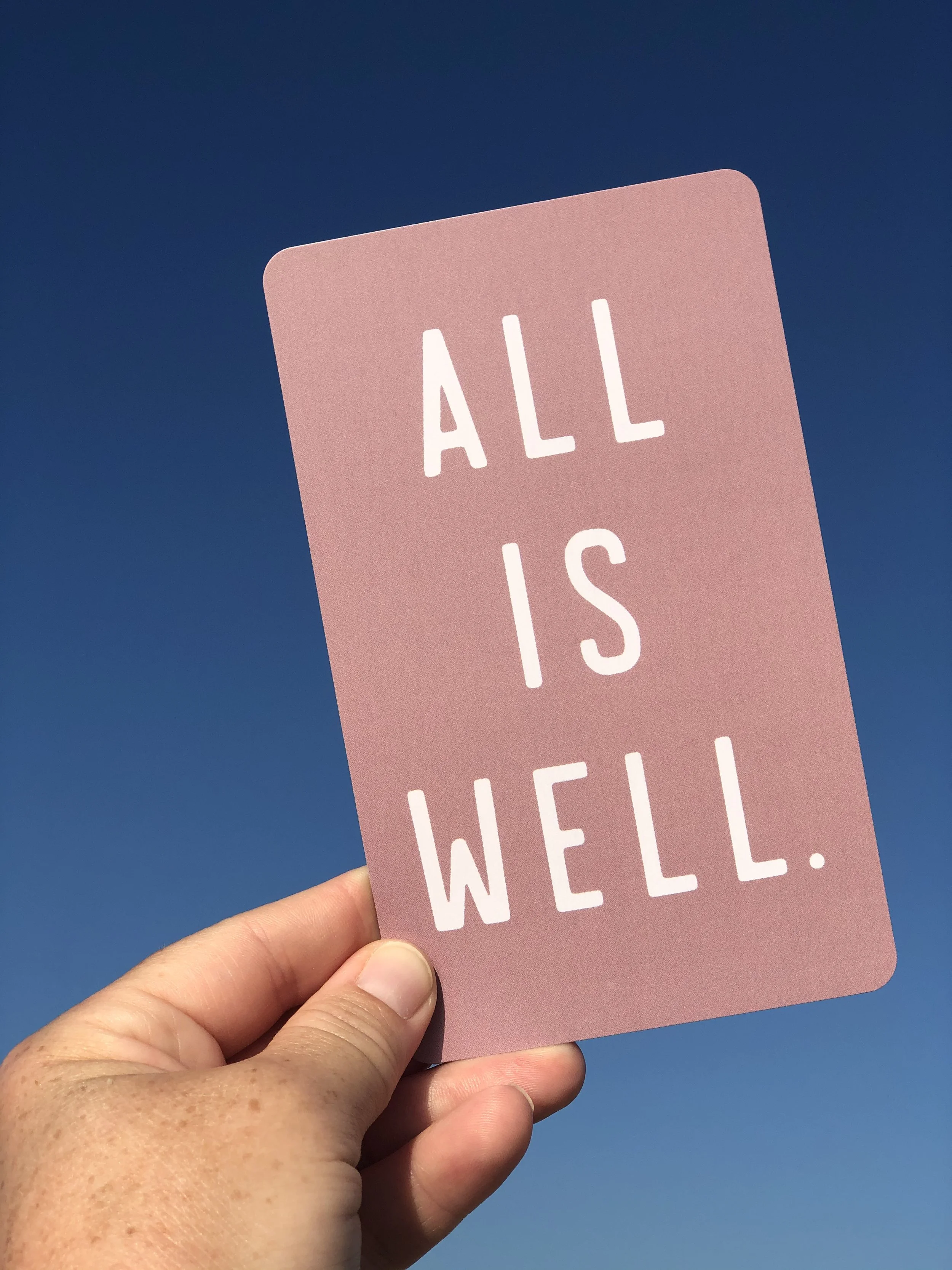 Close-up of a pink sign with white text reading 'ALL IS WELL' held up by a person's hand against a clear blue sky.