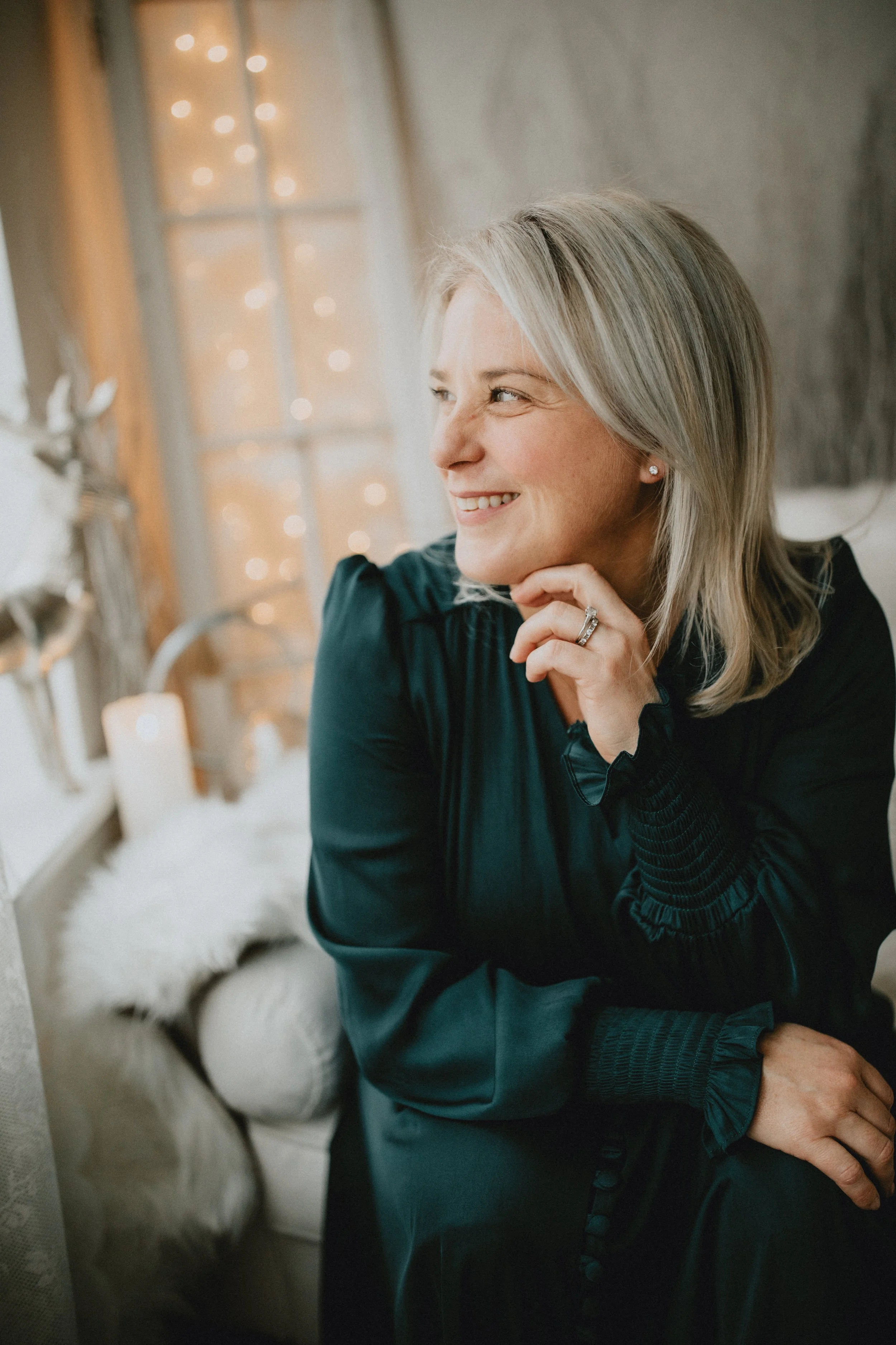 A smiling middle-aged woman with gray hair, wearing earrings and a dark green jacket, is resting her chin on her hand. She is indoors near a window with warm lighting and blurred Christmas lights in the background.