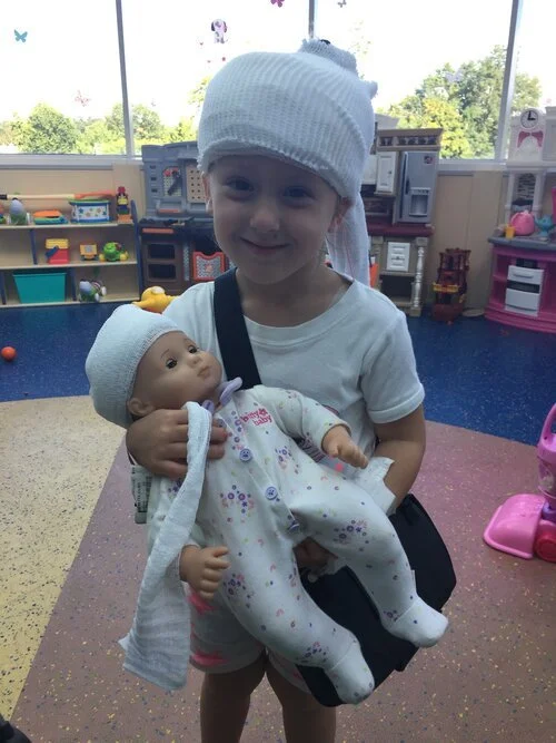 Young girl holding a baby doll in a playroom with toys and large windows in the background.
