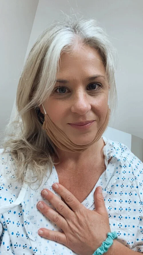 Woman with blonde hair in a hospital gown, wearing a face mask pulled down below her chin, touching her chest with her hand, smiling at the camera.