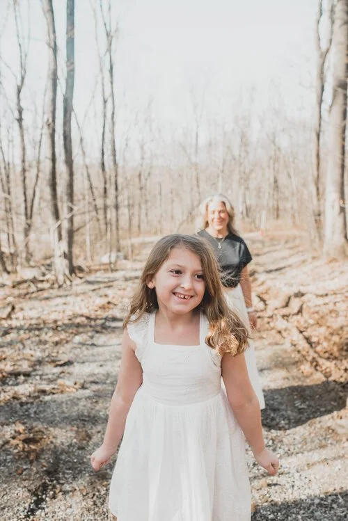 A young girl in a white dress smiling in front, with an older girl in a dark top and white skirt smiling in the background, walking on a wooded trail during daytime.