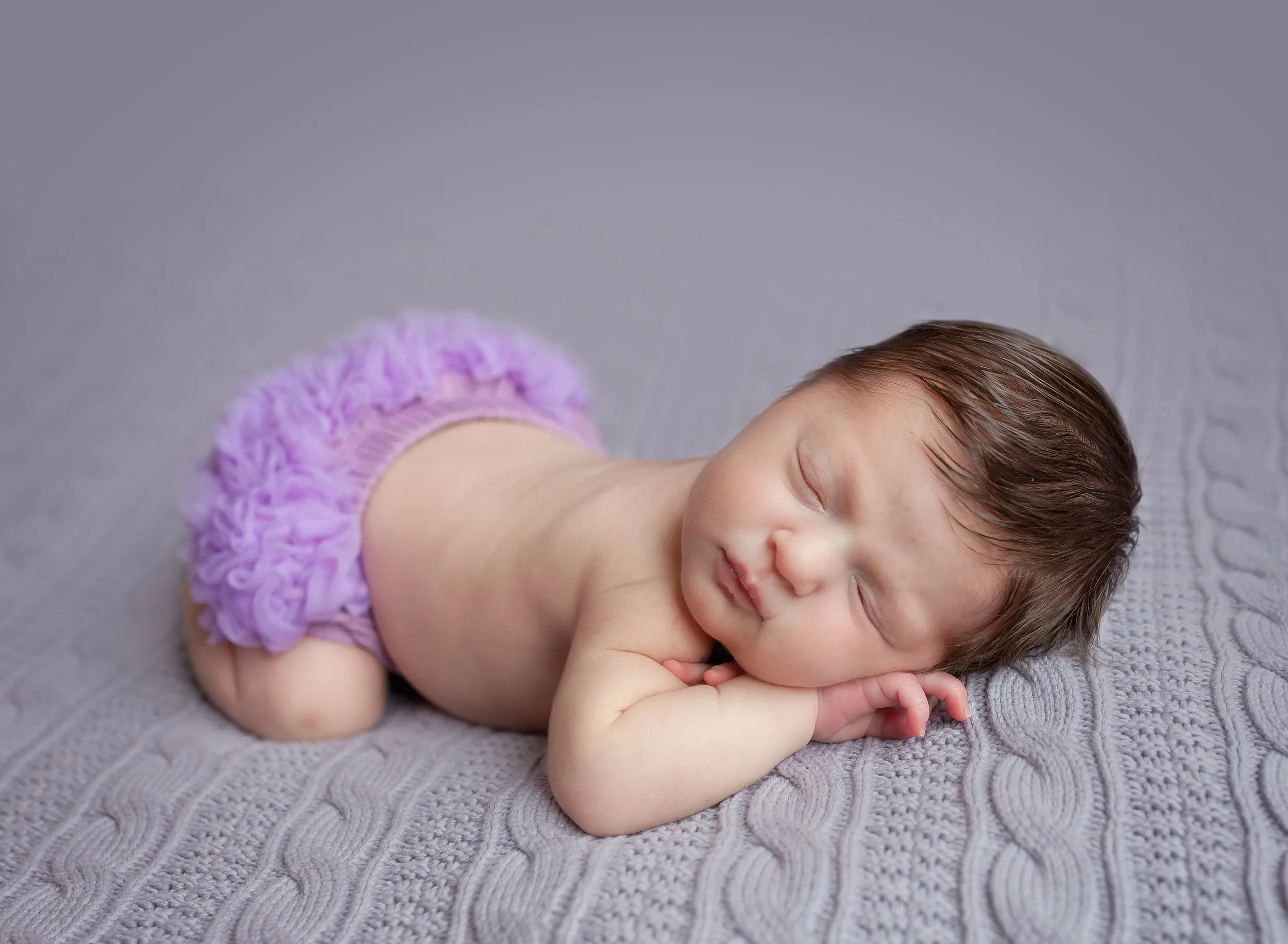 A sleeping baby with brown hair, lying on a textured gray blanket, wearing a purple ruffled diaper cover, resting with arms under the head on the blanket.
