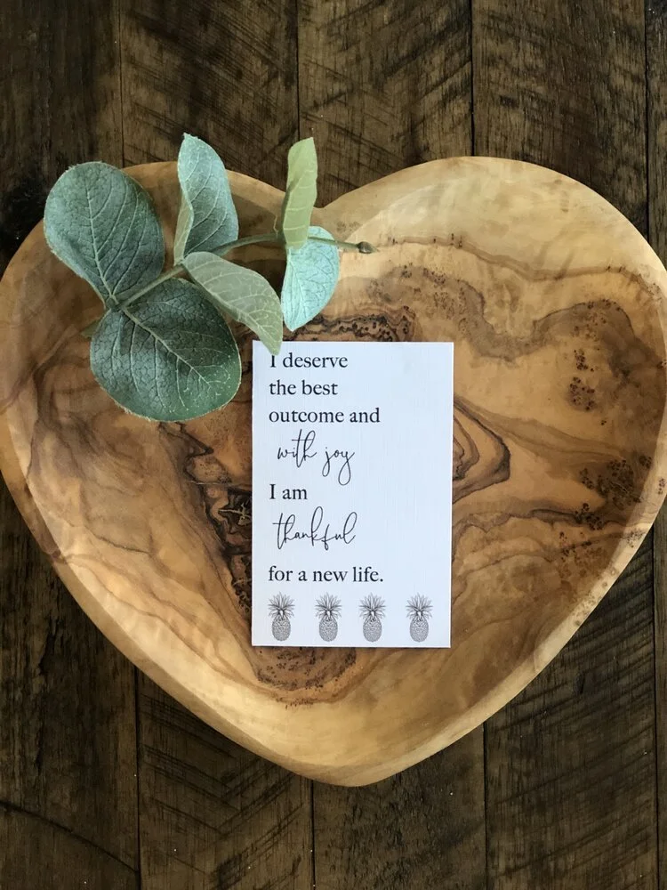 A wooden heart-shaped dish with a sprig of greenery and a motivational card that says, 'I deserve the best outcome and with joy I am thankful for a new life,' decorated with four small pineapples.
