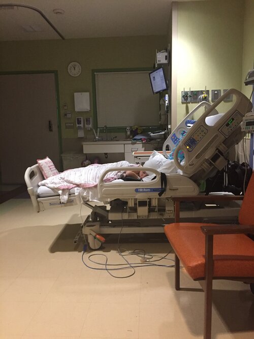 A hospital room with a patient lying in a hospital bed covered with a pink and white blanket. The bed is adjusted with the head raised and has side rails. There are medical monitors and equipment on the wall beside the bed, and a large window with a closed blind. An orange chair is positioned in front of the bed, and a clock on the wall shows the time as approximately 3:50. The room appears clean and organized.