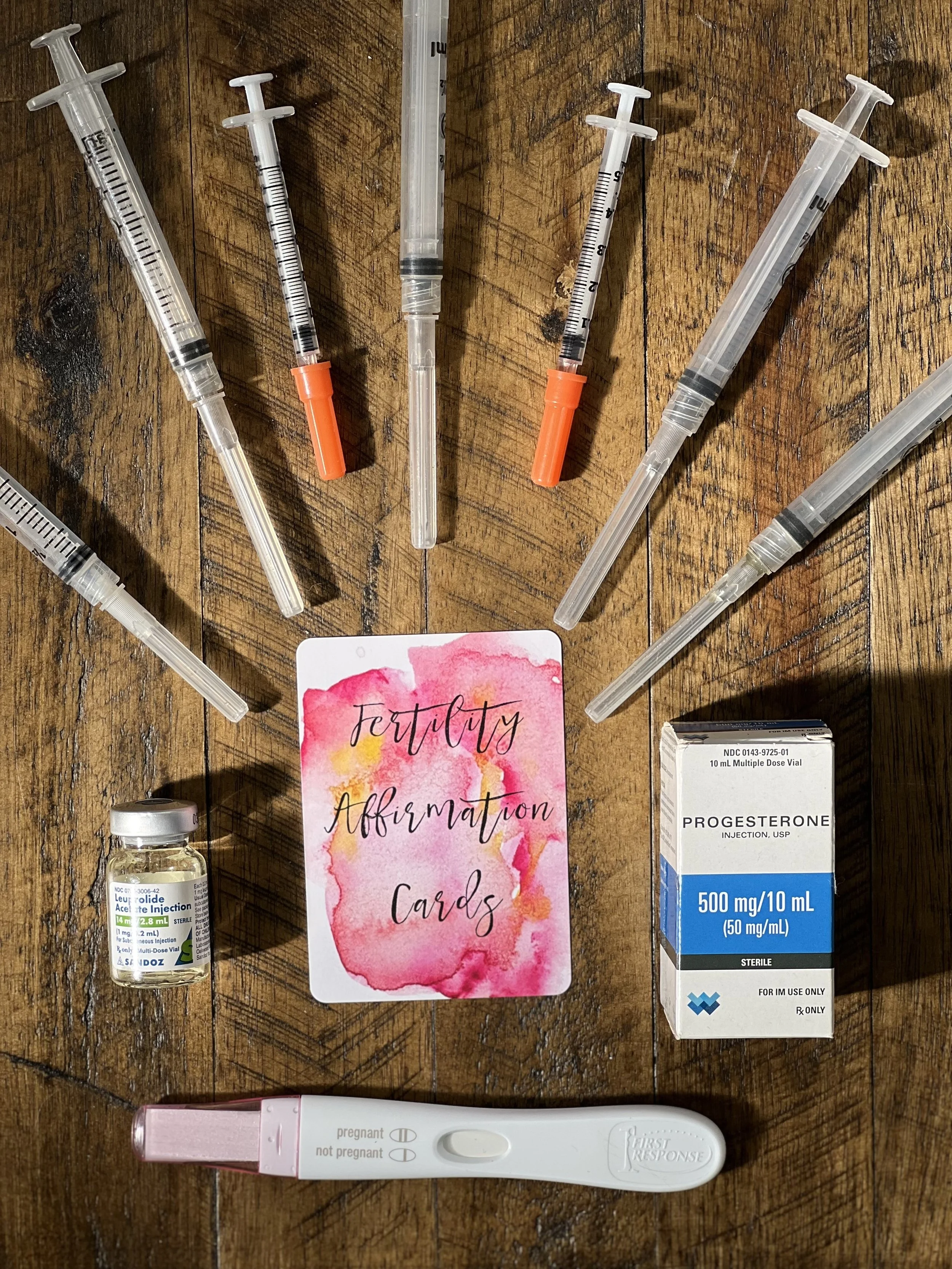 Several medical items on a wooden surface, including syringes, a box of progesterone injection, a pregnancy test, and a card that reads 'fertility affirmation cards'.