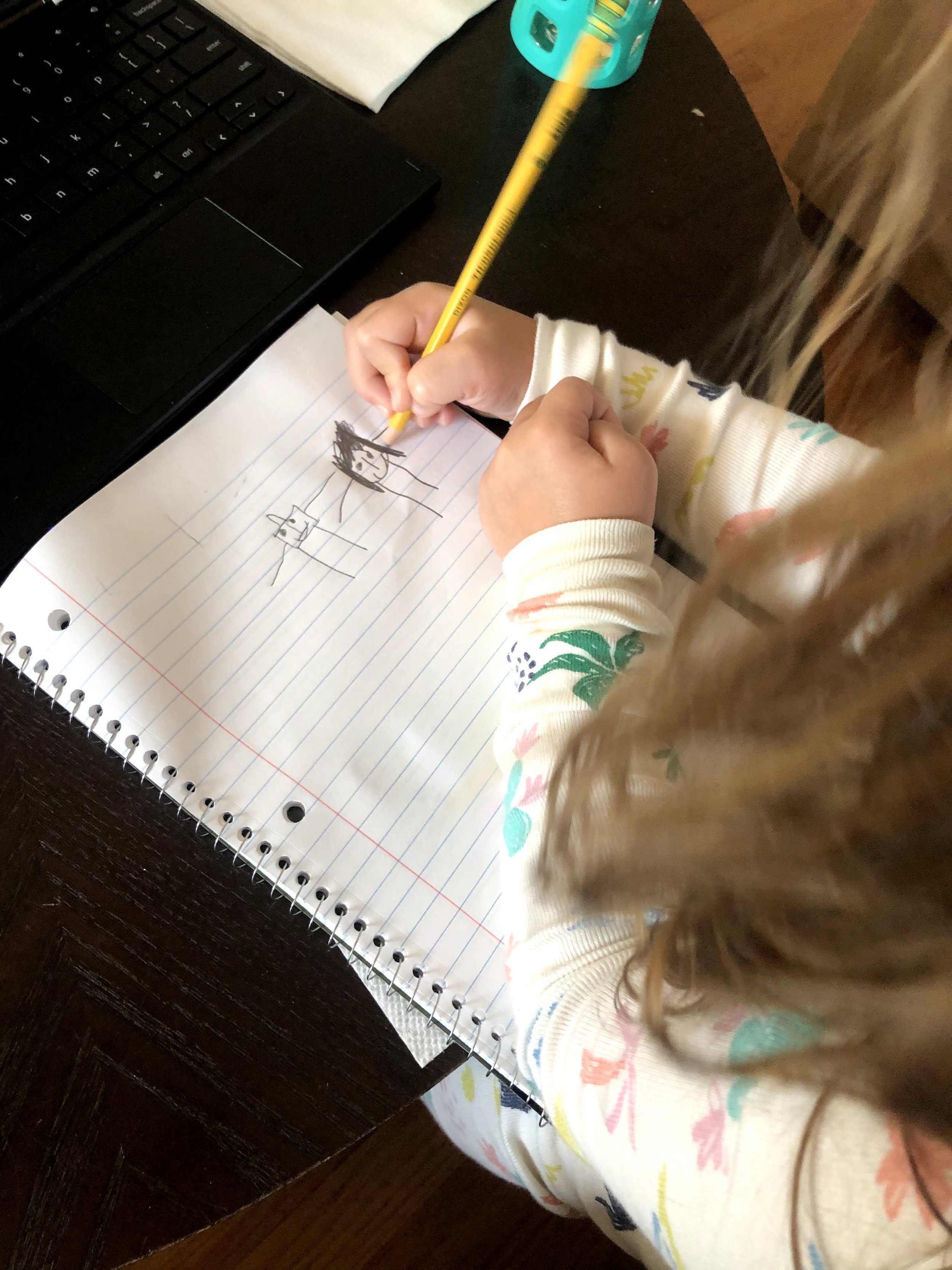 A child's hand drawing on lined notebook paper with a yellow pencil, with a laptop, a teal pencil holder, and a white napkin on a dark wooden table.