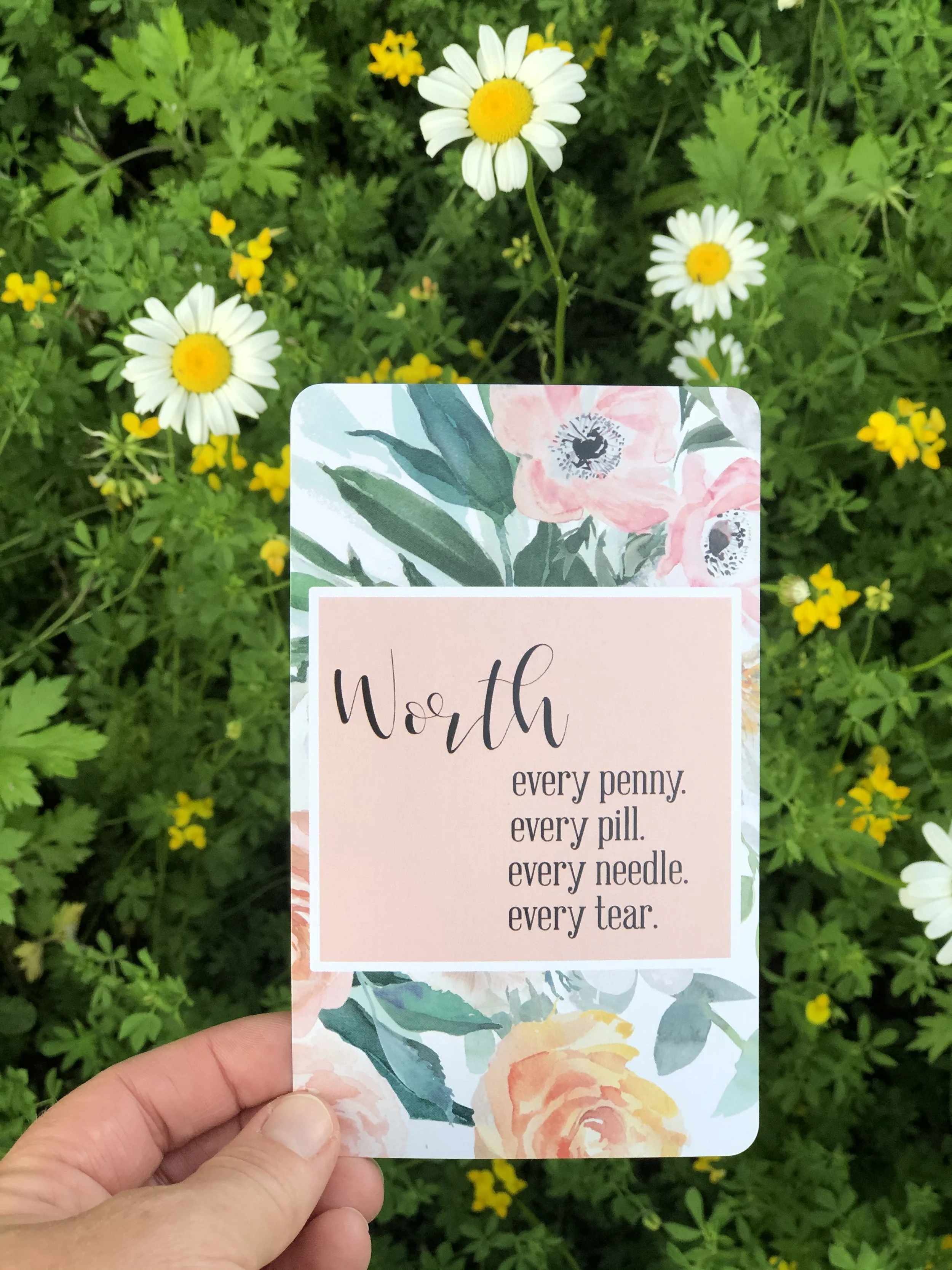 A hand holding a floral-themed card with the word "Worth" and an inspirational quote, set against a background of white daisies and yellow wildflowers.