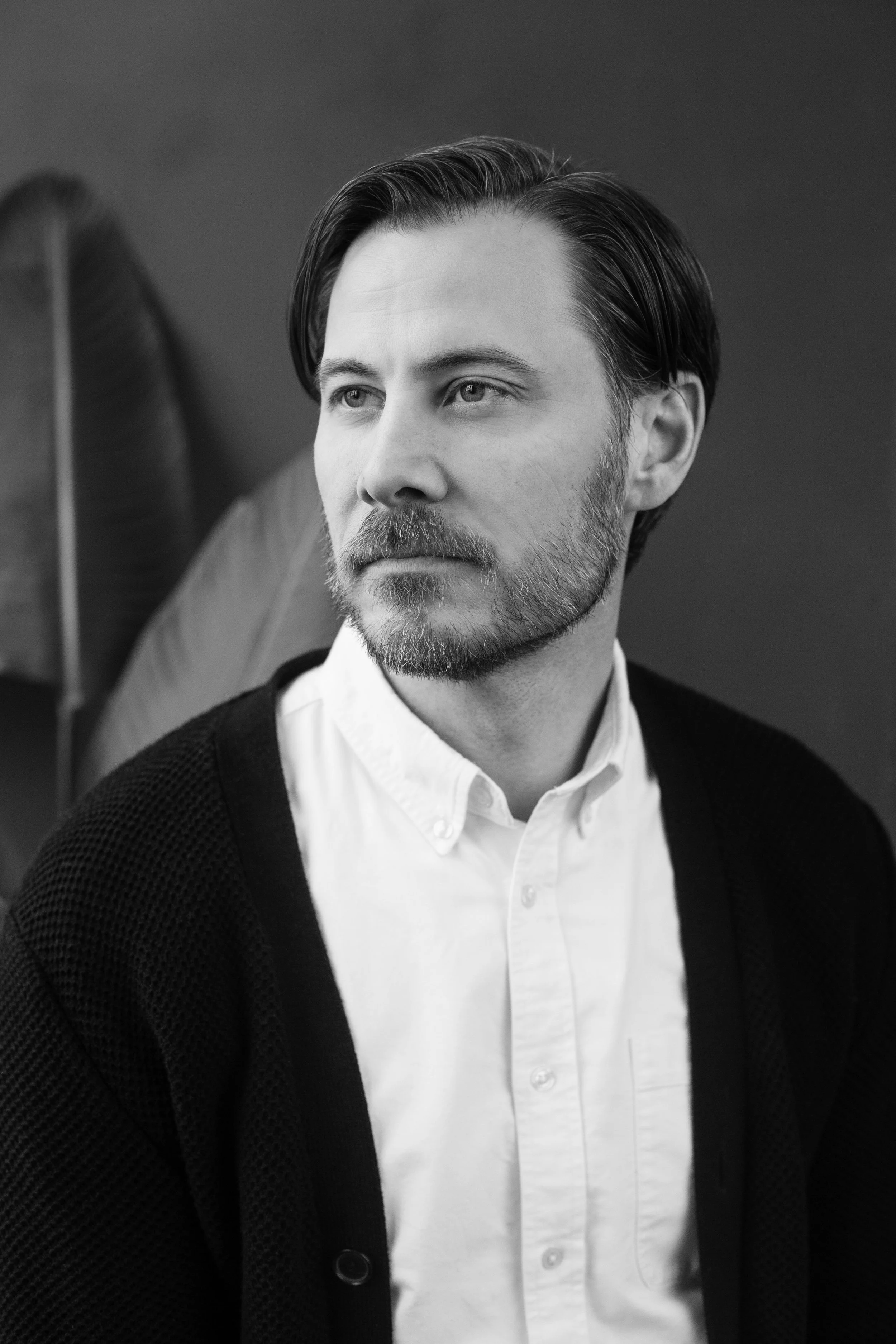Black and white portrait of a man with medium-length hair, a beard, wearing a white collared shirt and a dark cardigan, looking to the side.
