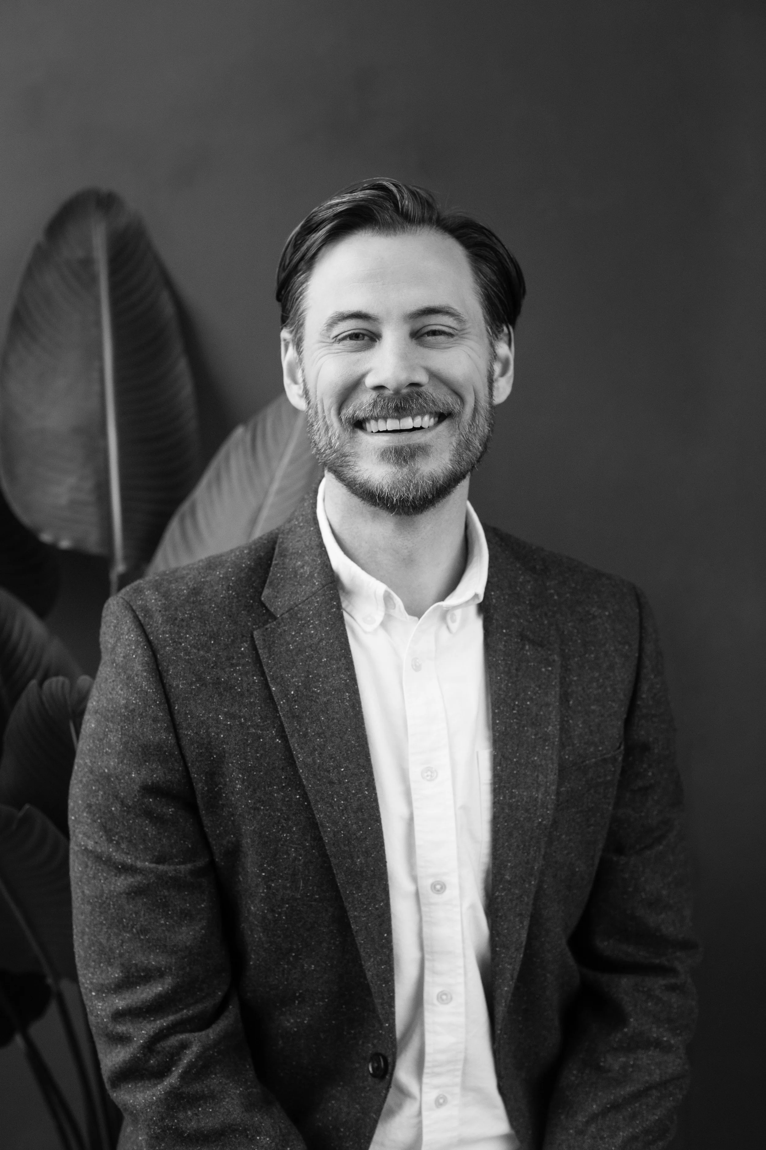 Black and white photo of a smiling man with a beard, wearing a suit jacket and white shirt, sitting in front of large leaves.