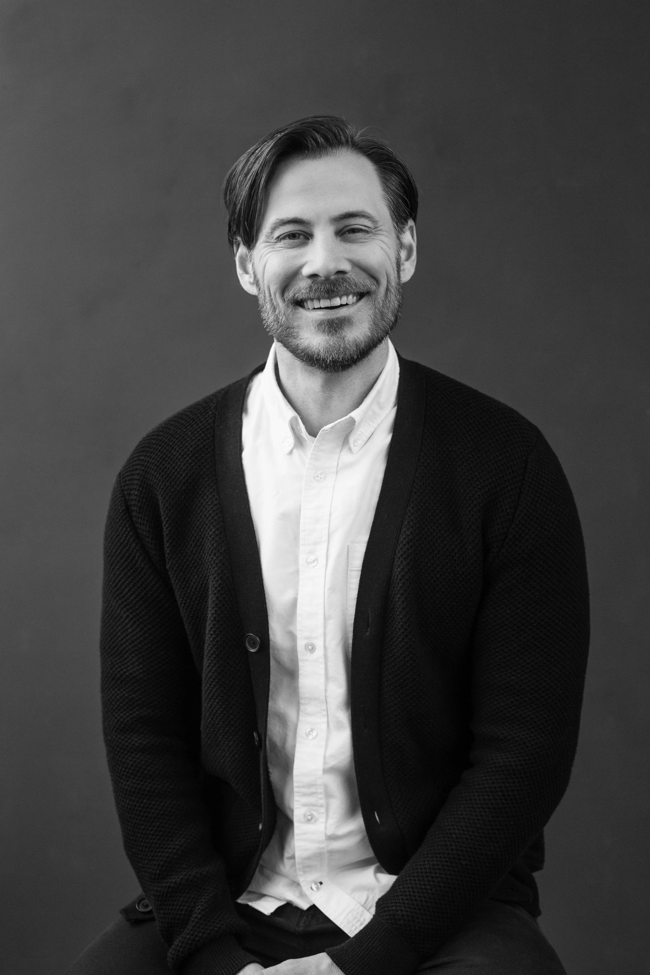 A man with a beard and medium-length hair smiling, wearing a white button-up shirt and a dark cardigan, sitting against a plain background.