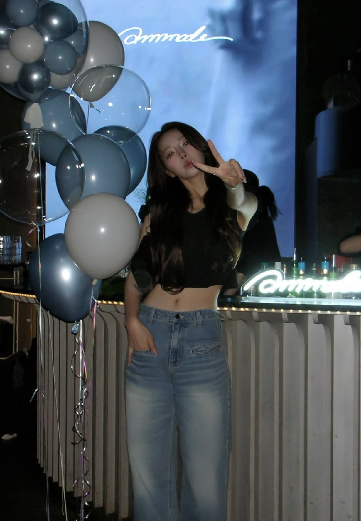 A young woman posing with a peace sign in front of a bar decorated with balloons and a neon sign at a celebration or party event.
