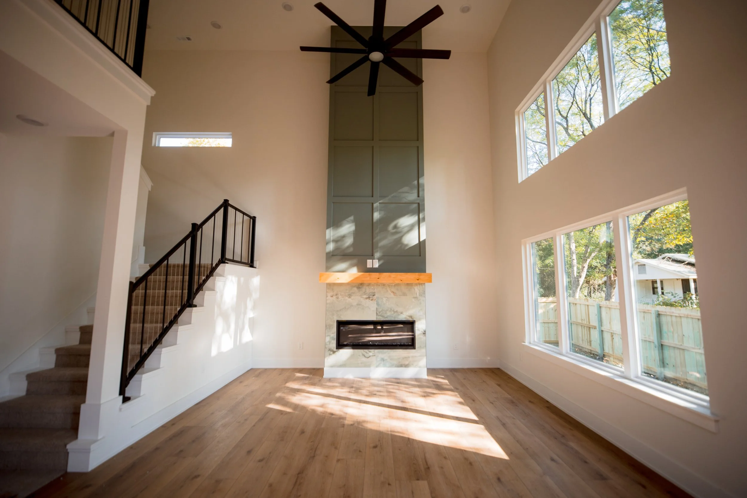 Empty living room with large windows, high ceiling, wooden floors, a modern fireplace with a marble surround, and a ceiling fan.