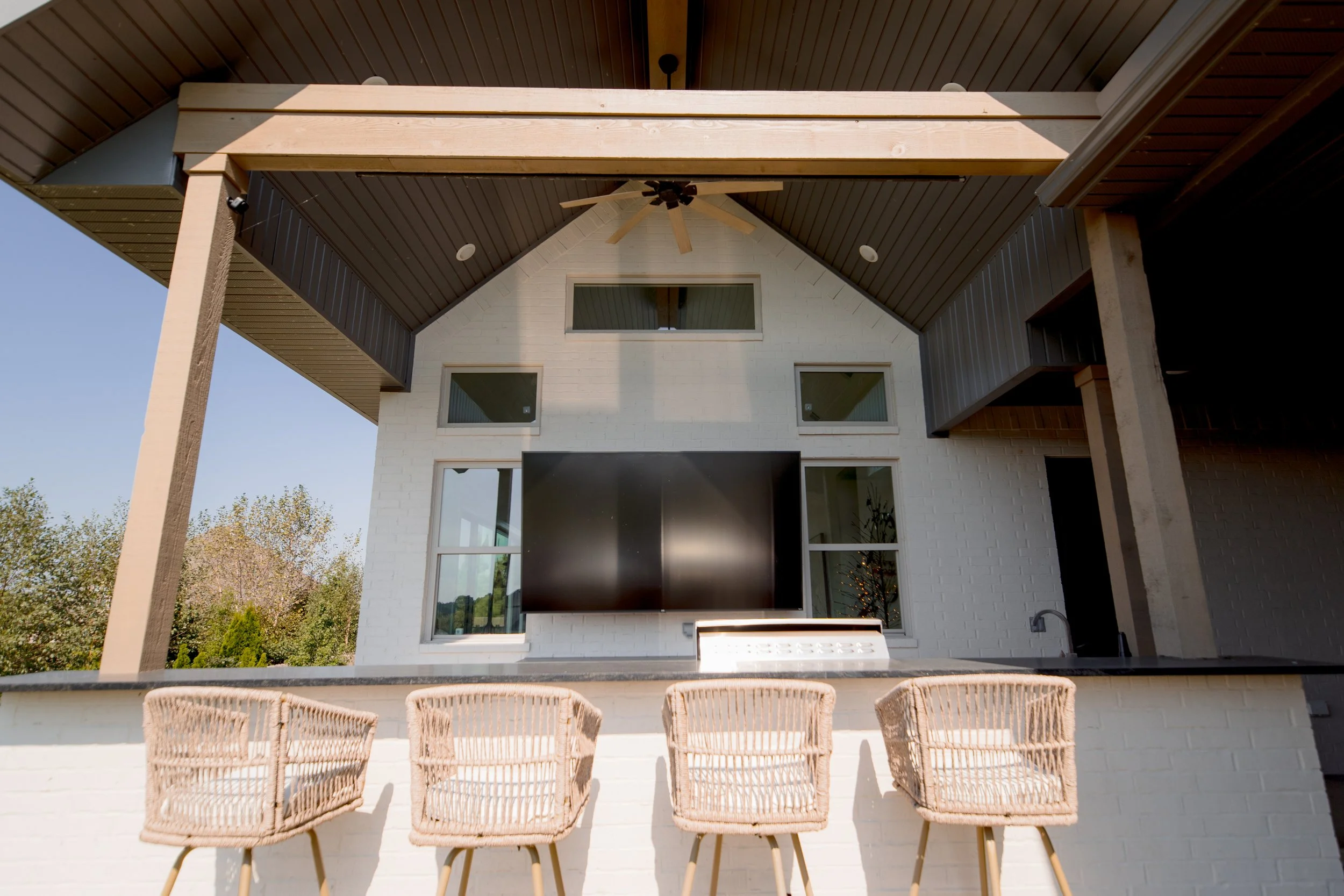 Modern outdoor patio with four wicker chairs facing a white brick wall and a large flat-screen TV, set under a wooden ceiling with a ceiling fan, with trees in the background.