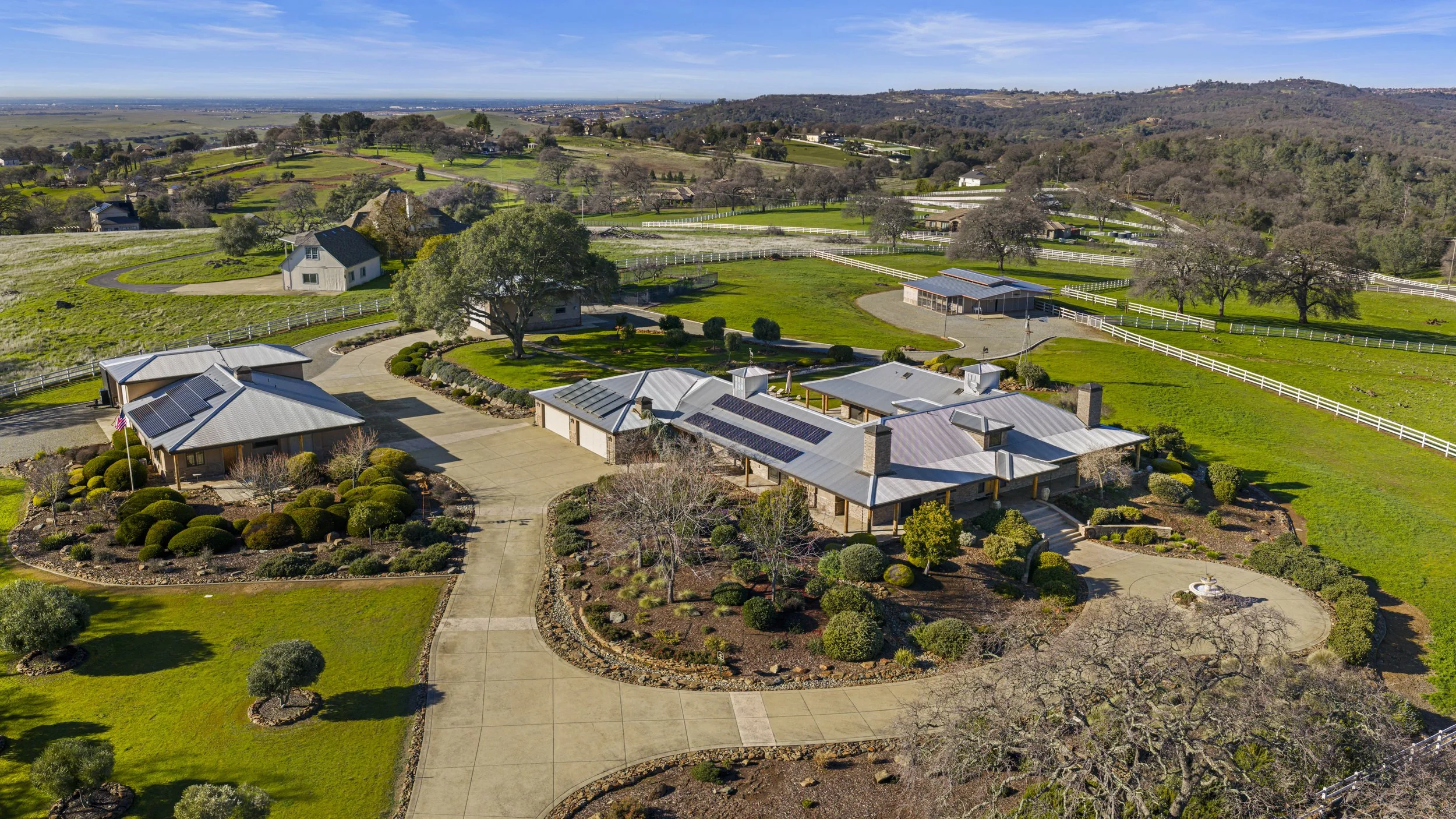 Aerial view of a large countryside property with a main house, landscaped gardens, outbuildings, and rolling green pastures surrounded by white fencing.