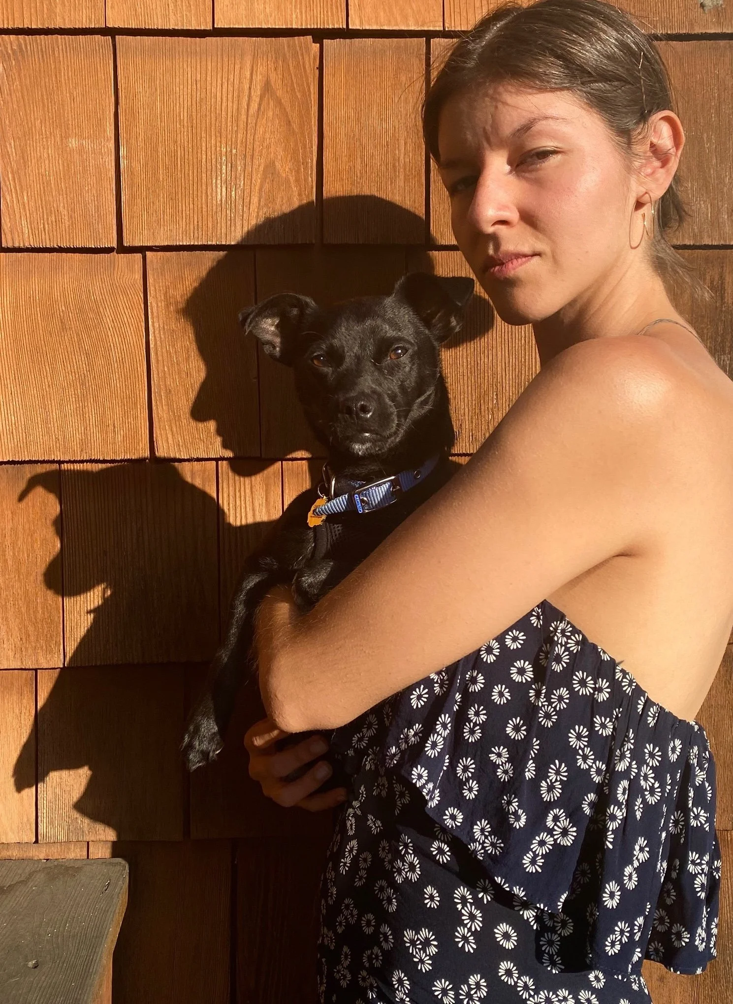 woman holds small dog in arms against a shingled wall
