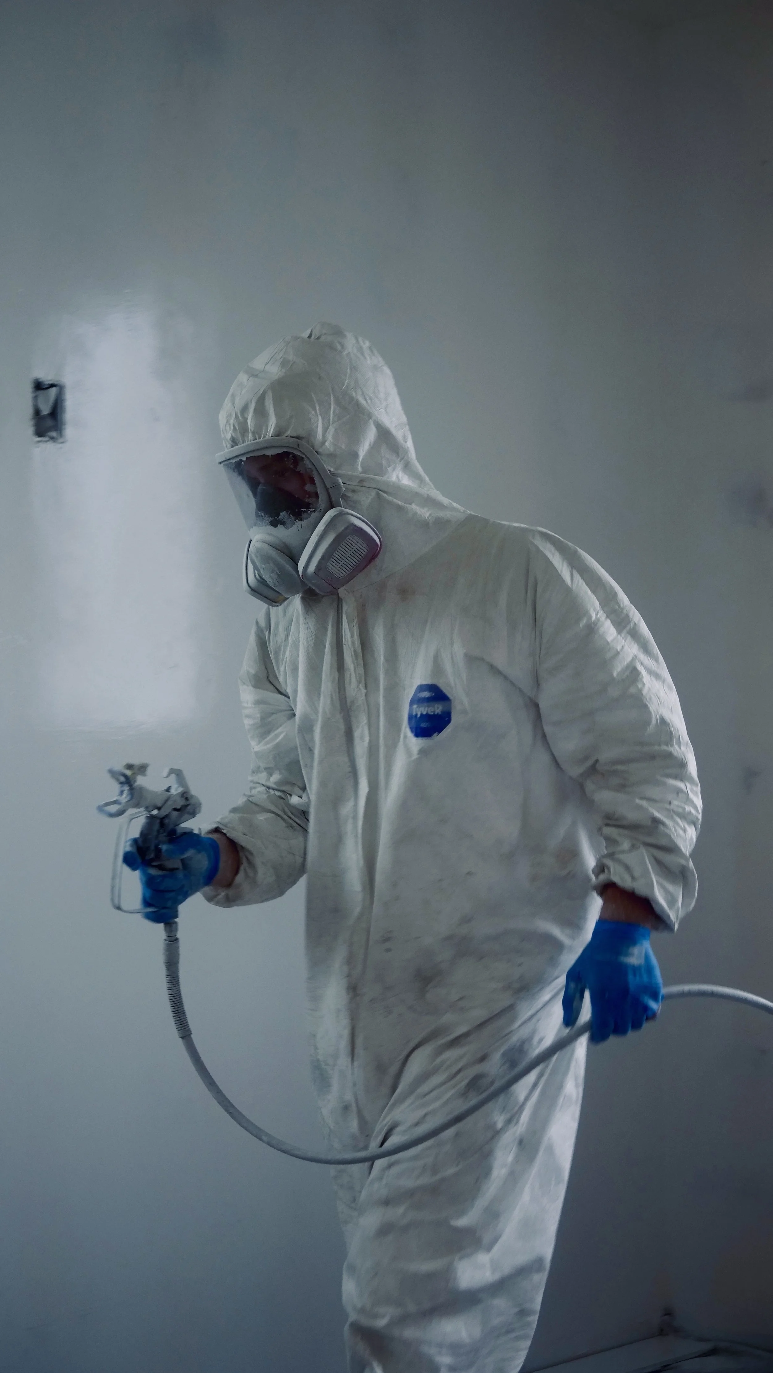 A person dressed in full protective white hazmat suit, headset with a mask, and blue gloves, holding a spray nozzle, working in a hazmat environment.