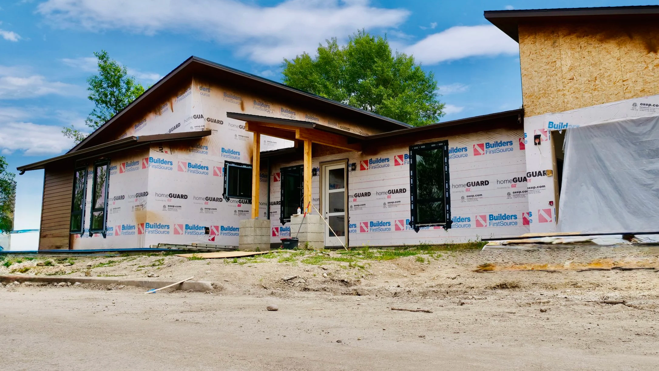 A house under construction with partially finished walls and windows, with a clear blue sky and green trees in the background.