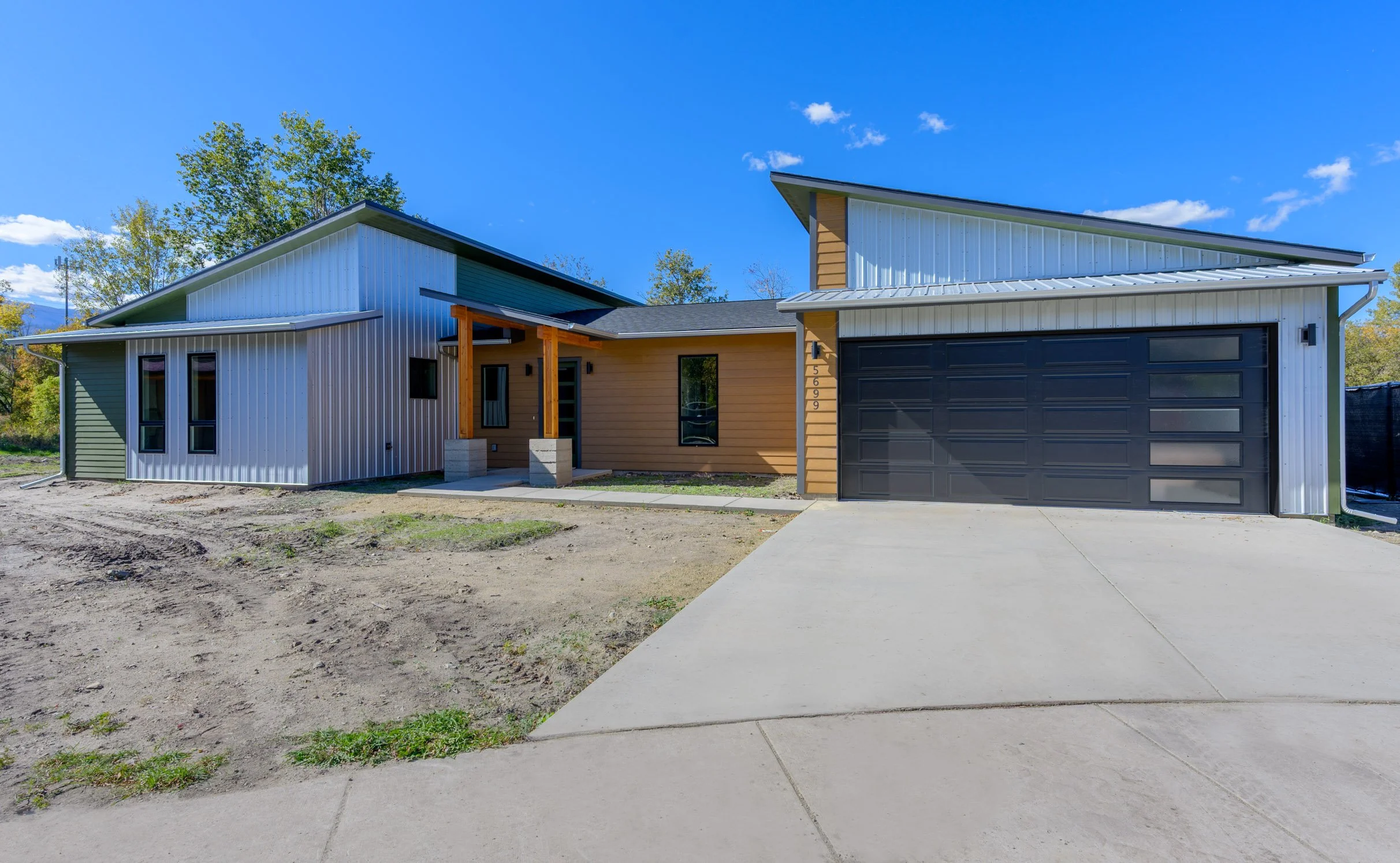 Newly constructed modern house with a combination of metal and wood siding, a two-car garage with blackout panels, and a concrete driveway against a backdrop of trees and a bright blue sky.