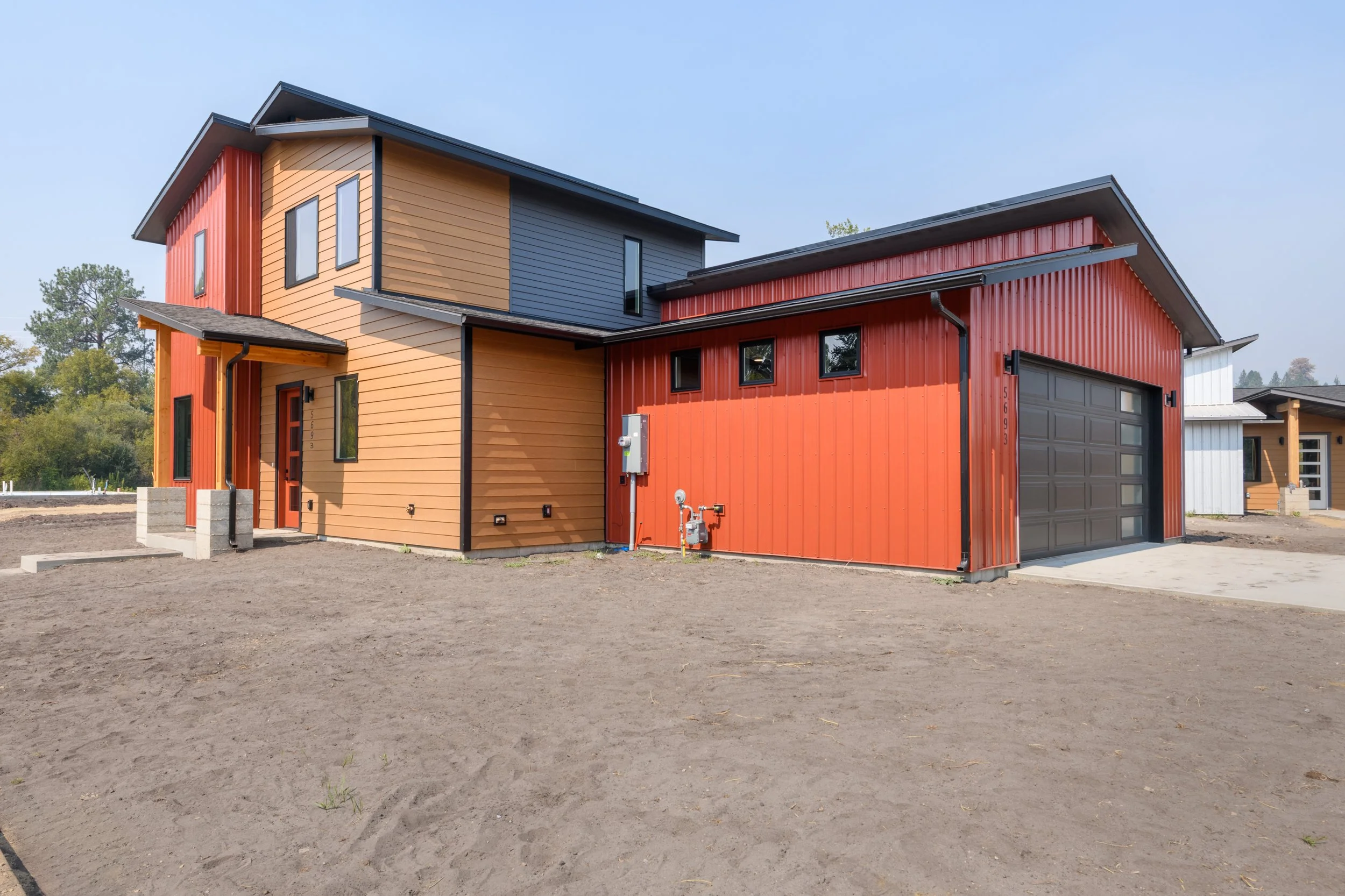 A modern two-story house with a colorful exterior, including red, yellow, blue, and white paneling. The house has multiple windows, a garage door, and is situated on a large plot with a dirt foreground and trees in the background.