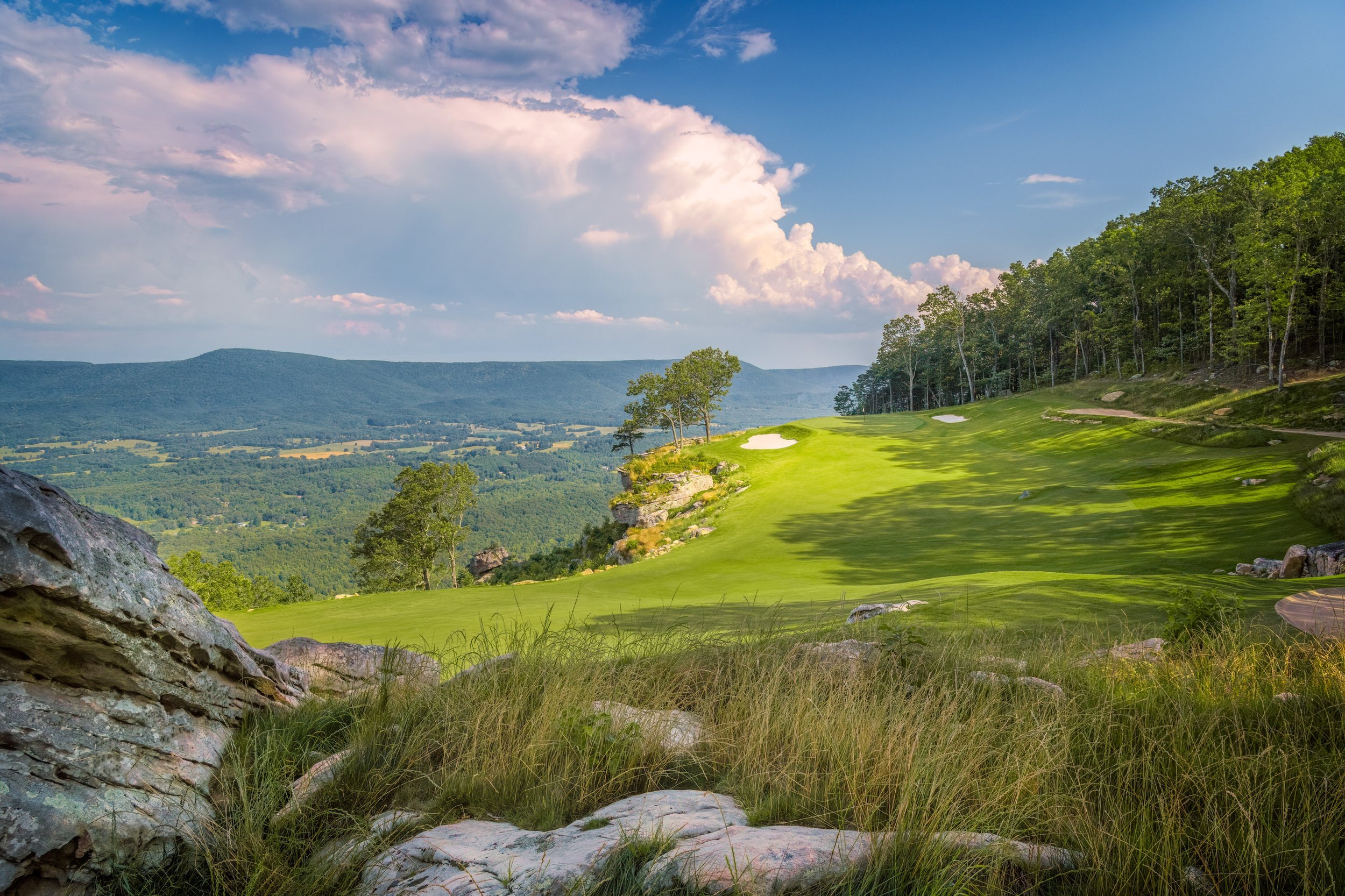 A scenic view of a golf course on a hillside with lush green grass, trees, and a blue sky with clouds.