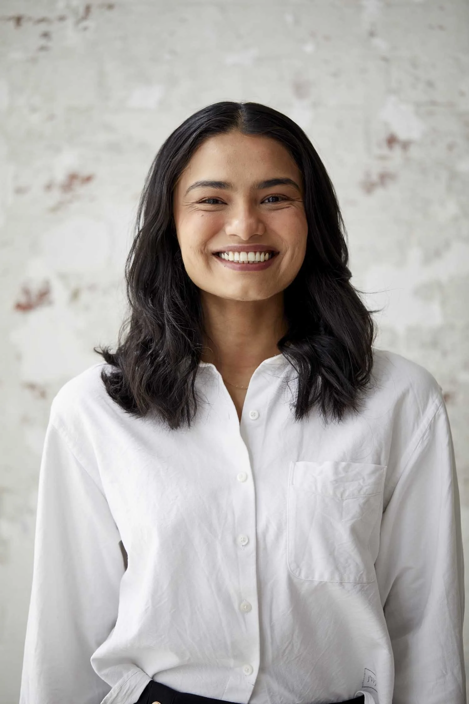 A woman with dark, wavy hair and a light smile, wearing a white button-up shirt, standing against a light-colored, slightly textured wall.