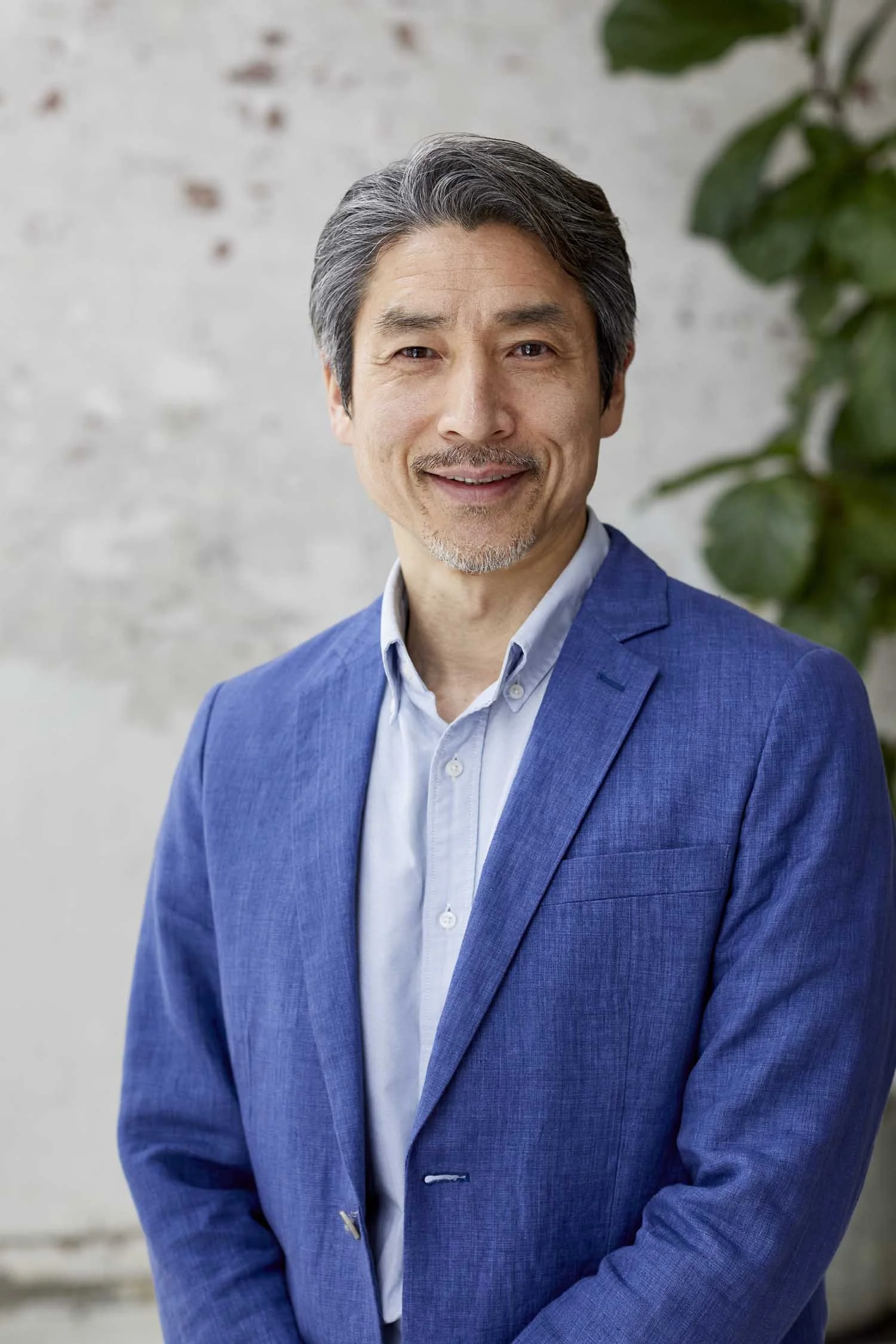 A middle-aged man with salt and pepper hair and a goatee, wearing a blue suit jacket over a light blue shirt, smiling softly against a light-colored wall with green foliage in the background.