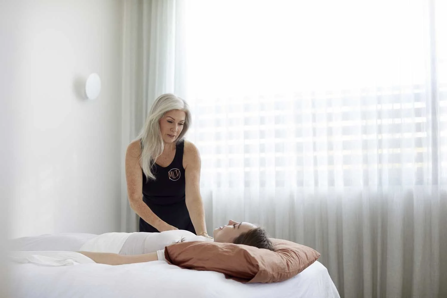 A woman with long blonde hair in a black sleeveless top tending to a young girl lying on a bed with a white sheet and brown pillow, in a bright room with white curtains.