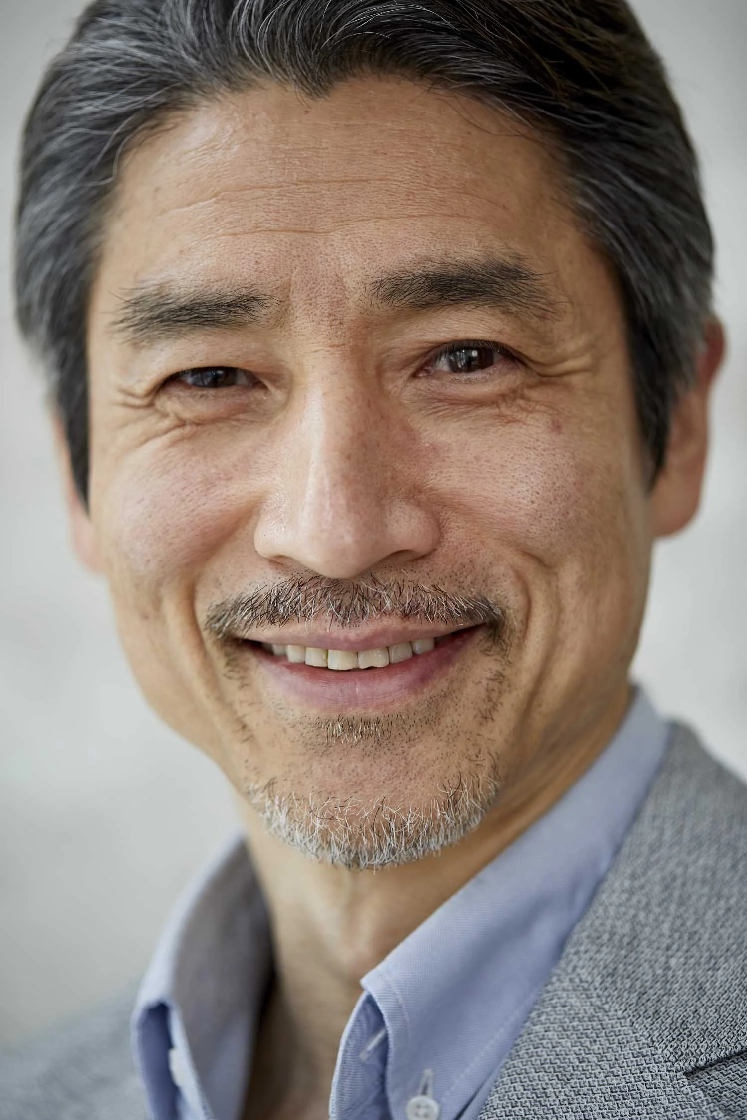 Close-up portrait of a smiling middle-aged East Asian man with gray hair and a gray beard, wearing a light blue shirt and gray blazer.