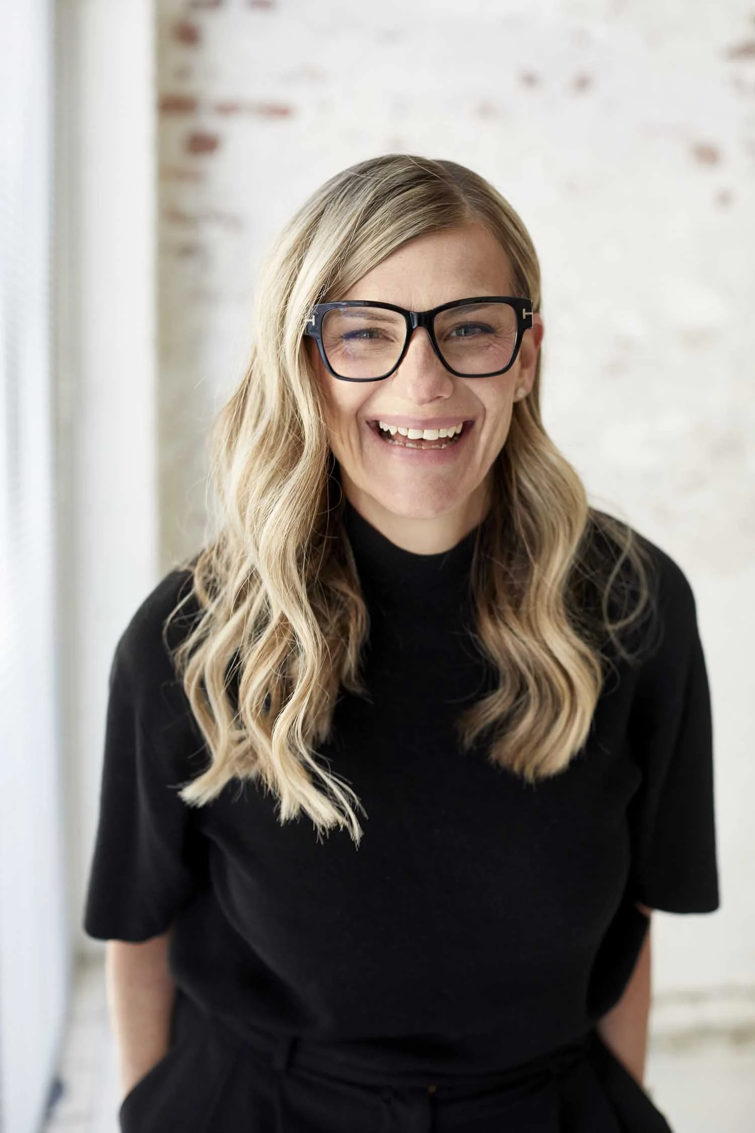 A woman with long wavy blonde hair, wearing black glasses and a black top, smiling and looking at the camera, with a blurred background of a white wall with exposed brick details.
