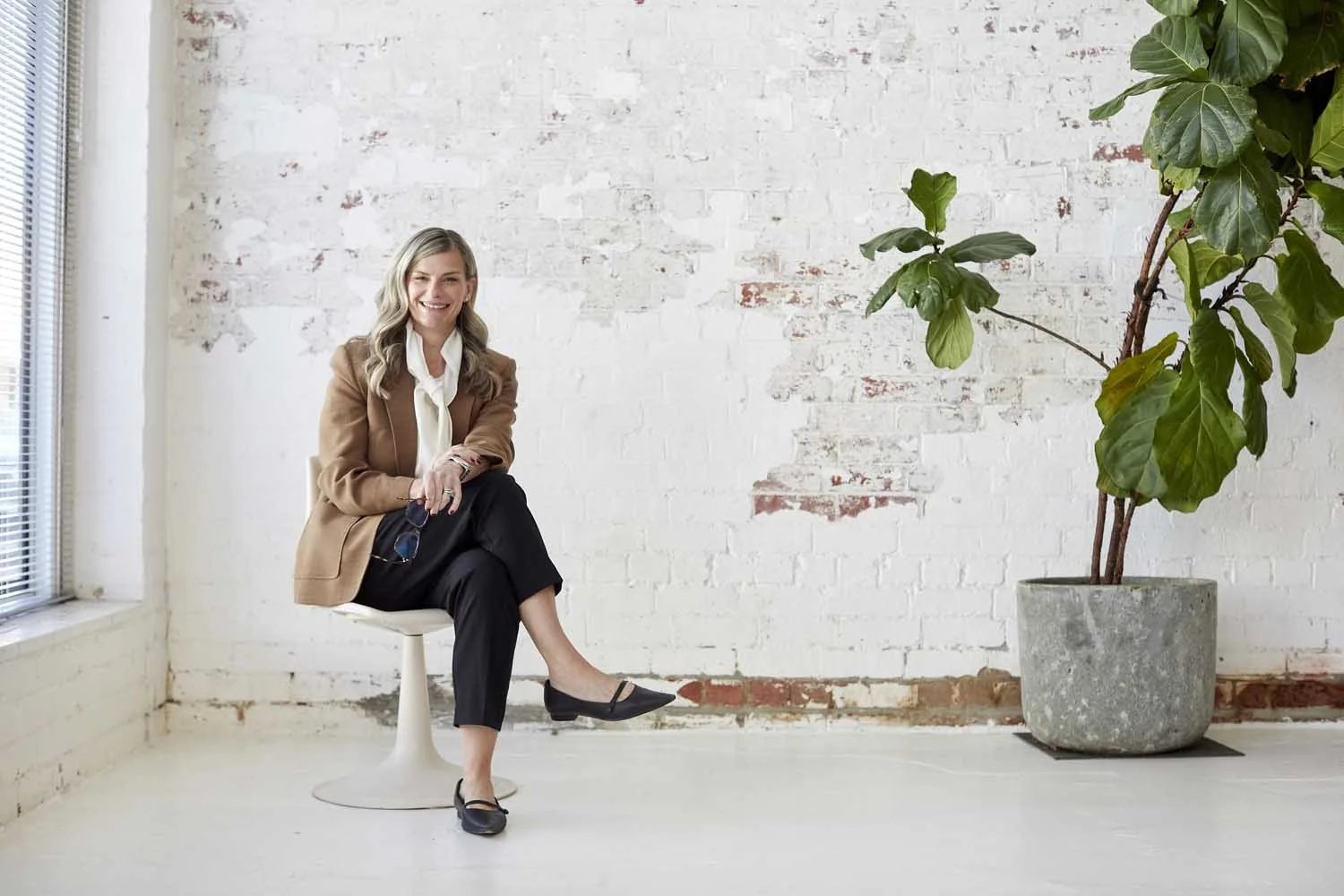 A woman with blonde hair wearing a tan blazer, white blouse, and black pants sitting on a white stool in a room with white brick walls and a large potted plant.