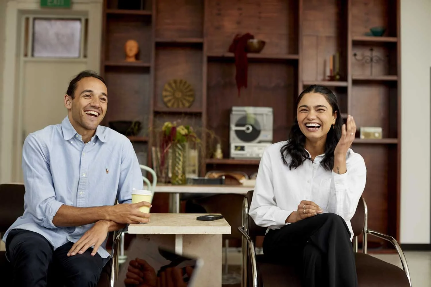 Two people sitting on chairs at a table, laughing, in a cozy room with wooden shelves and eclectic decor.