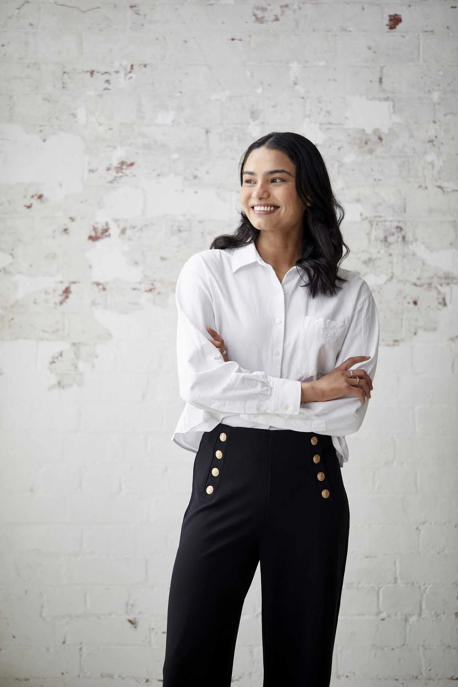 A woman with dark hair in loose waves, wearing a white button-up shirt and black pants with gold buttons, standing with arms crossed in front of a white brick wall with some red paint marks, smiling to the side.